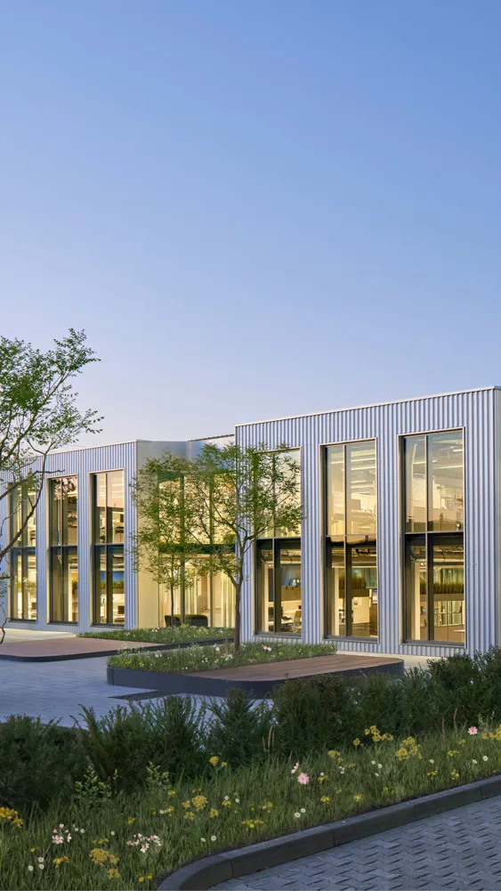 Modernes Bürogebäude mit großen Fenstern, Bäumen, Blumen und gepflastertem Parkplatz bei Sonnenuntergang.