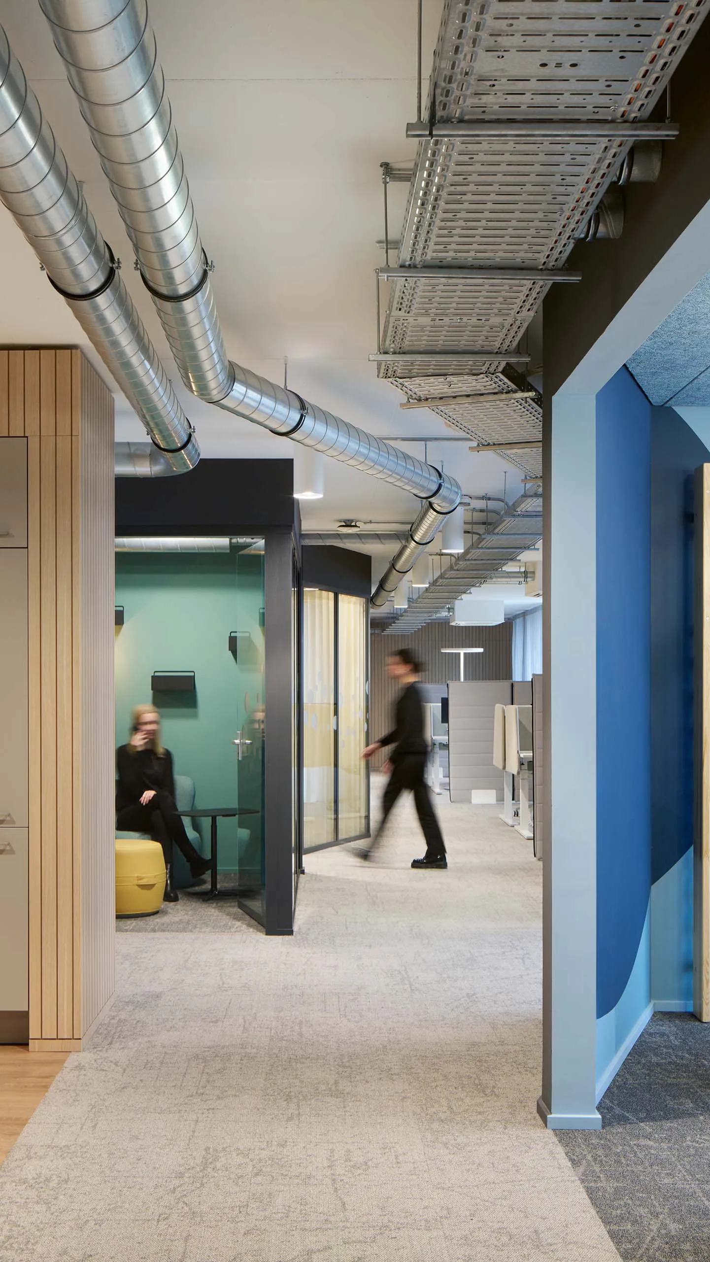 Modernes Büro mit offenen Arbeitsbereichen, Holzverkleidungen, sichtbaren Lüftungsrohren und blauen Wandelementen mit Fitnessgeräten.