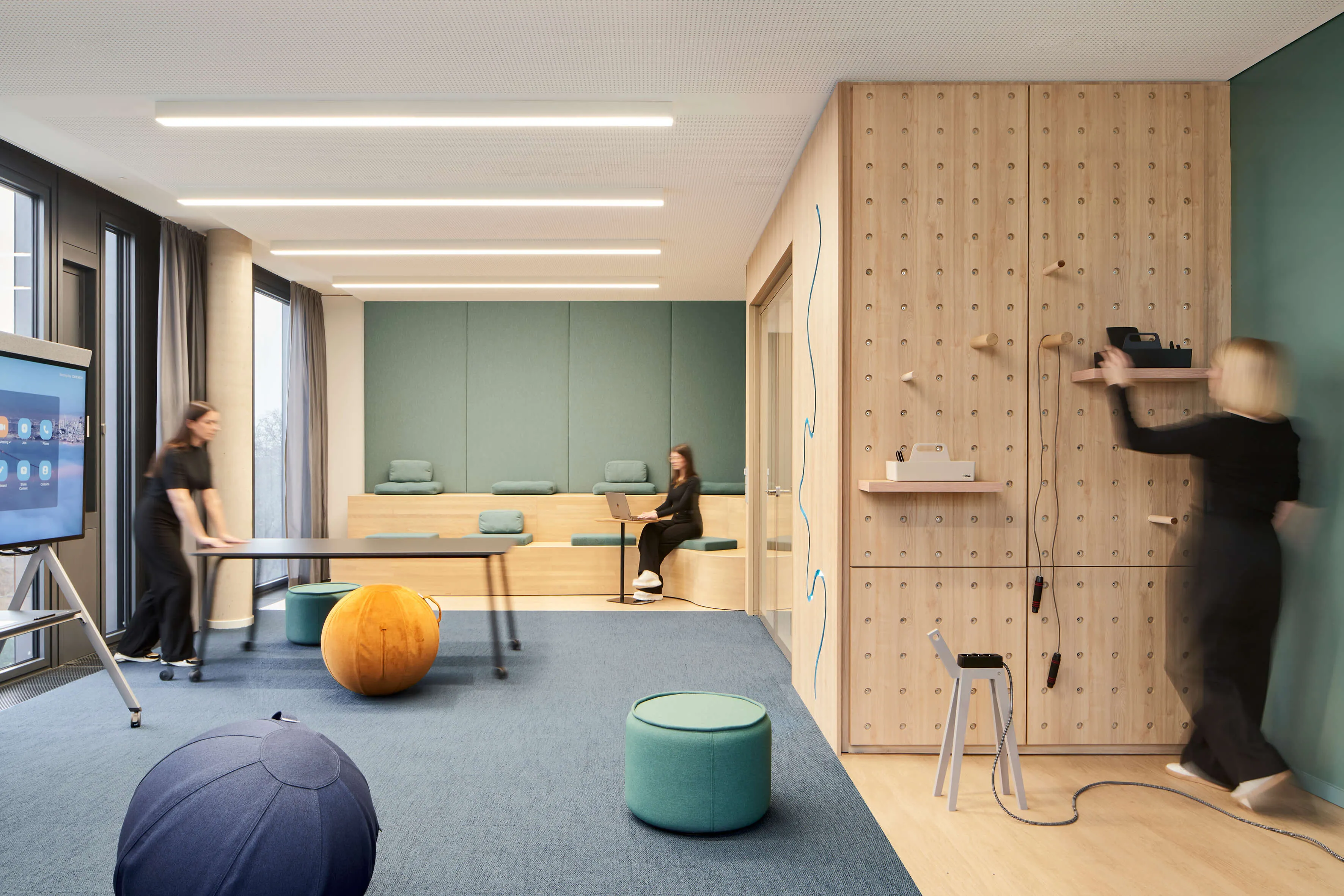 Modernes Büro mit blauem Teppich, Holzwand mit Klettergriff und Sitzinsel, zwei Frauen arbeiten und richten einen Tisch her.