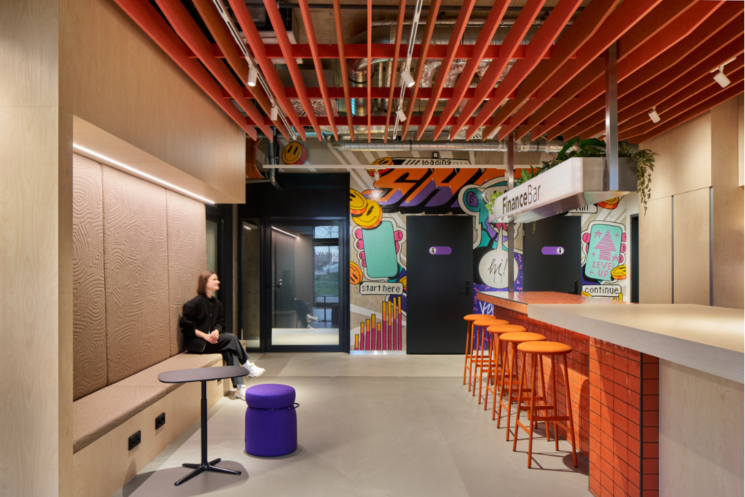 Moderner Innenraum mit bunten Barhockern, Wandgrafiken und einer Frau, die auf einer gepolsterten Bank sitzt.