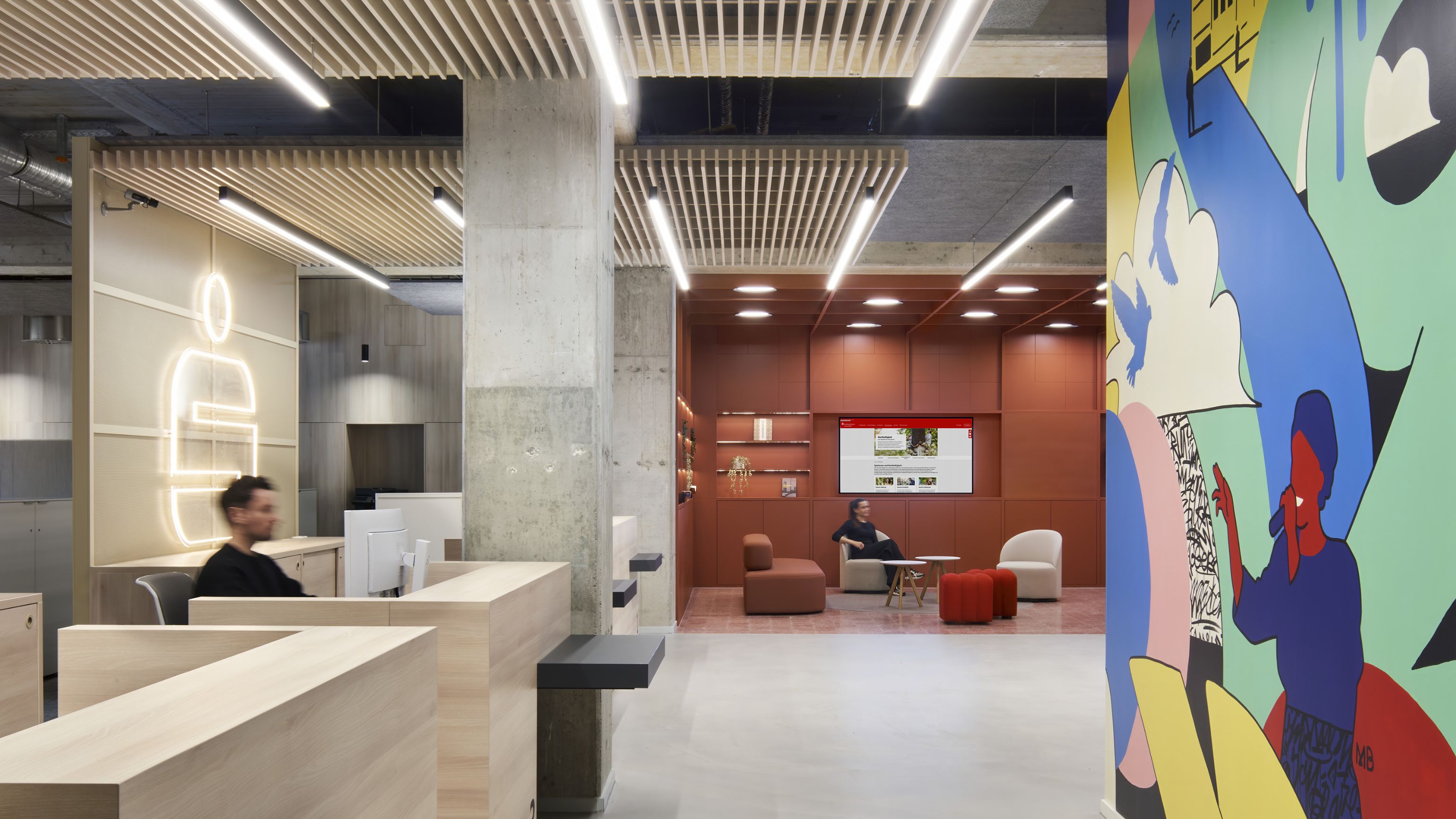 Moderner Empfangsbereich mit Holztresen, Betonpfeilern und warmem Lichtakzent – im Hintergrund eine terrakottafarbene Lounge-Zone mit Sesseln und Digital-Signage, rechts ein farbenfroher Wandmural.