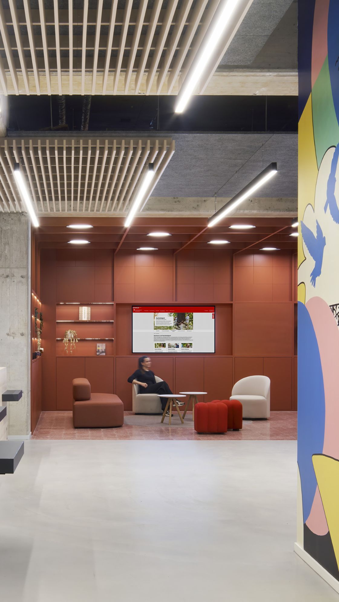 Moderner Empfangsbereich mit Holztresen, Betonpfeilern und warmem Lichtakzent – im Hintergrund eine terrakottafarbene Lounge-Zone mit Sesseln und Digital-Signage, rechts ein farbenfroher Wandmural.