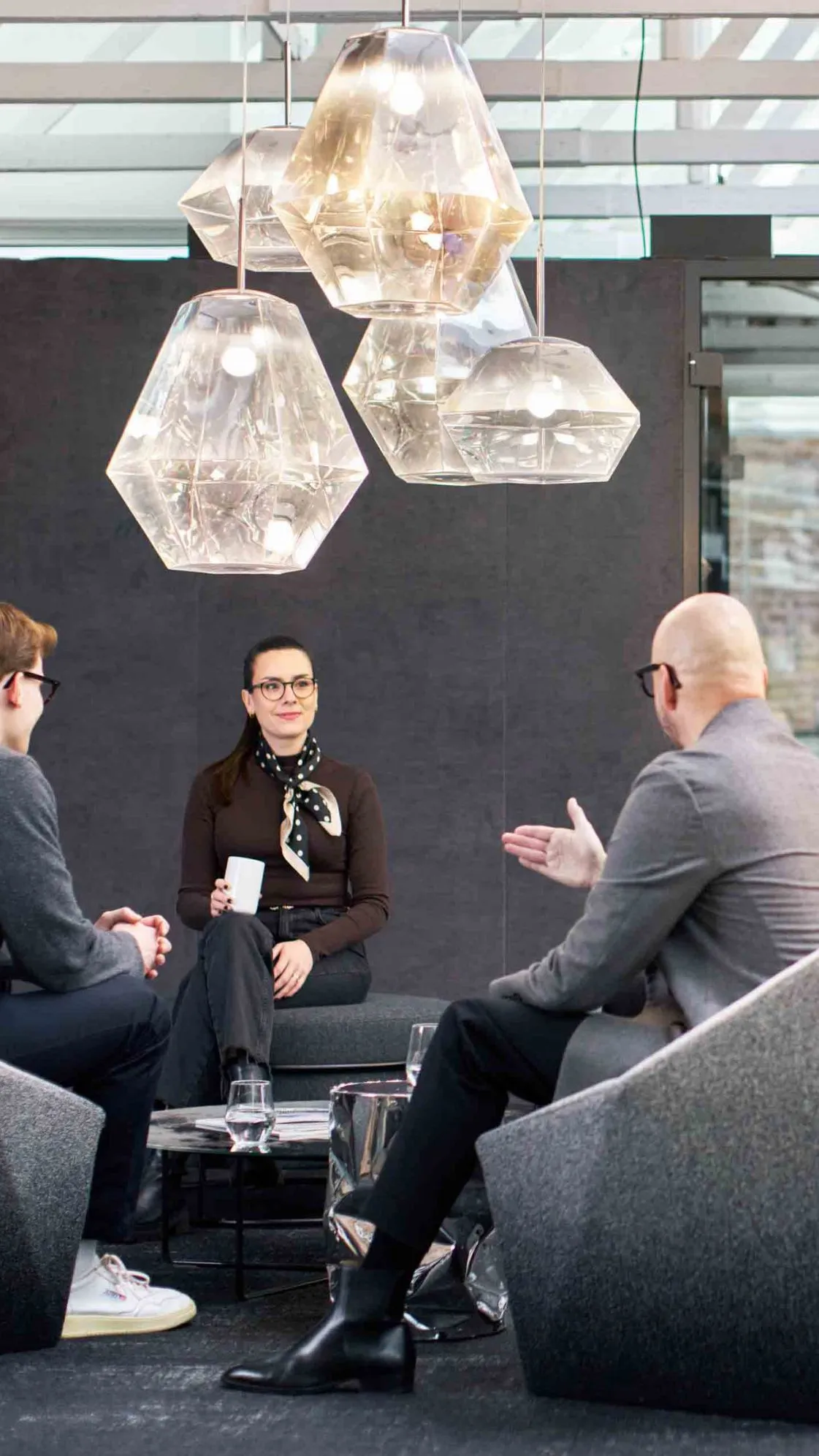Drei Personen im Gespräch auf modernen Lounge-Sesseln in einem offen gestalteten Büro – unter markanten Pendelleuchten aus Glas, mit Sichtmauerwerk und transparenten Raumteilern im Hintergrund.