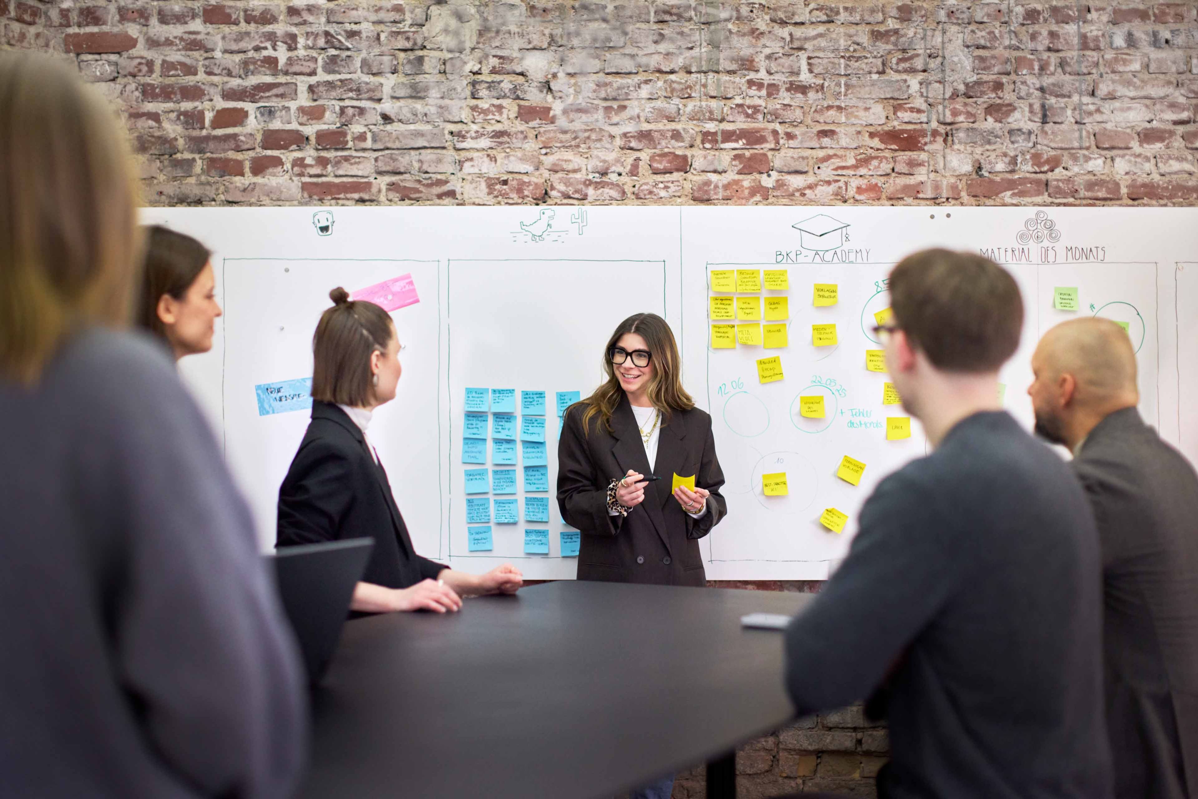 Teammeeting bei bkp: Marie Seliger präsentiert vor einem großen Whiteboard mit bunten Post-its und Skizzen, Teilnehmende sitzen am Konferenztisch vor Sichtmauerwerk.
