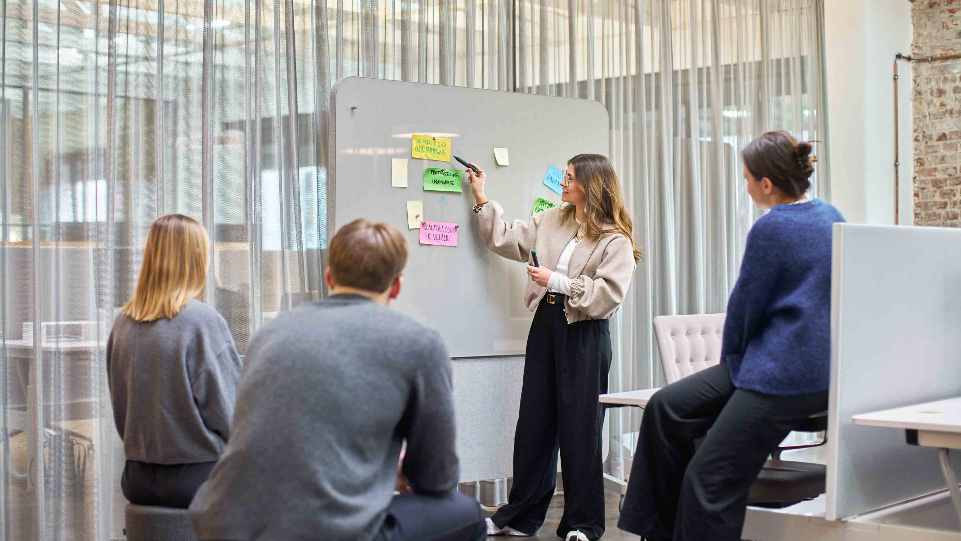 Team-Workshop im bkp-Büro: Marie Seliger am Glasboard mit Post-its, drei aufmerksame Zuhörende im lichtdurchfluteten Raum.