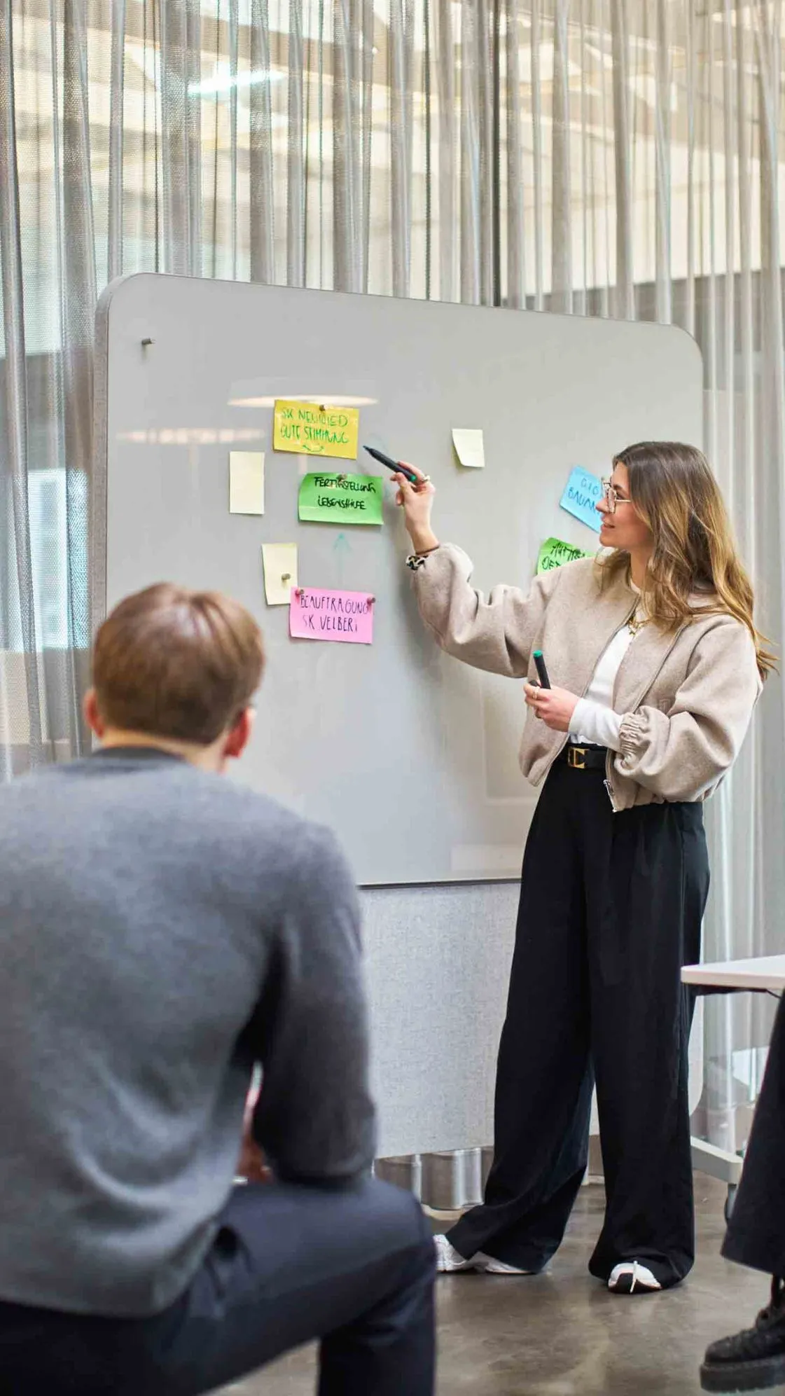 Team-Workshop im bkp-Büro: Marie Seliger am Glasboard mit Post-its, drei aufmerksame Zuhörende im lichtdurchfluteten Raum.
