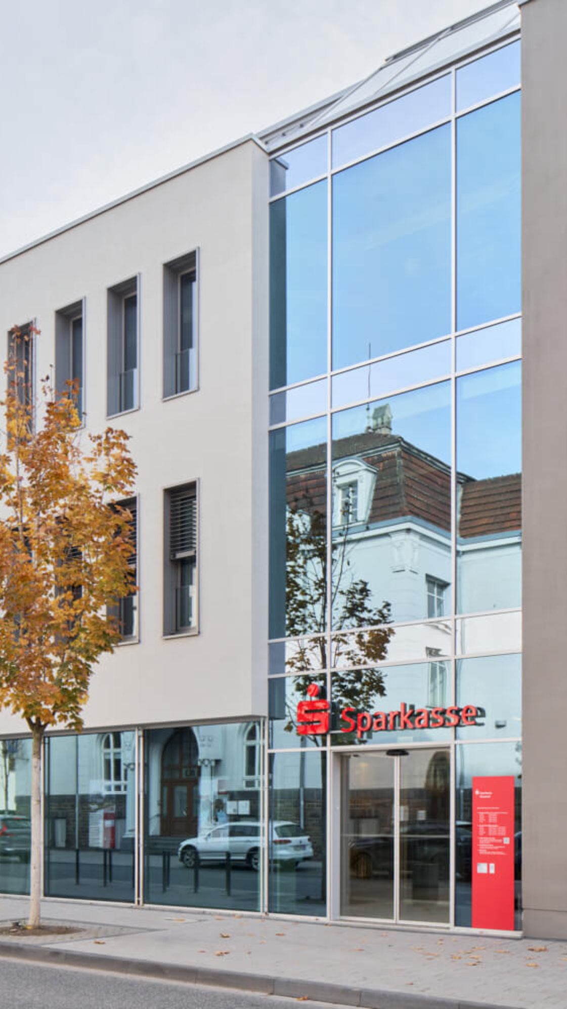 Außenansicht der Sparkasse-Filiale mit moderner Glasfront, rotem Sparkasse-Logo und herbstlichem Straßenbaum im Vordergrund.