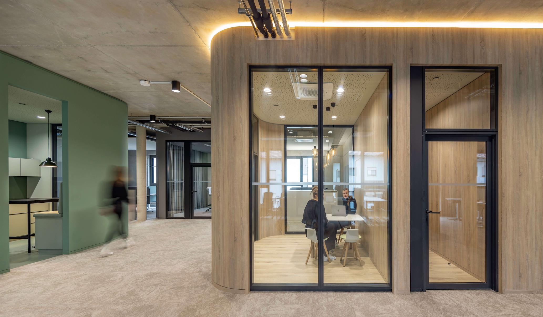 Modernes Büro mit verglastem Besprechungspod aus Holz: Zwei Personen im vertraulichen Meeting am Laptop, im Hintergrund offene Bürofläche mit grünen Akzenten.