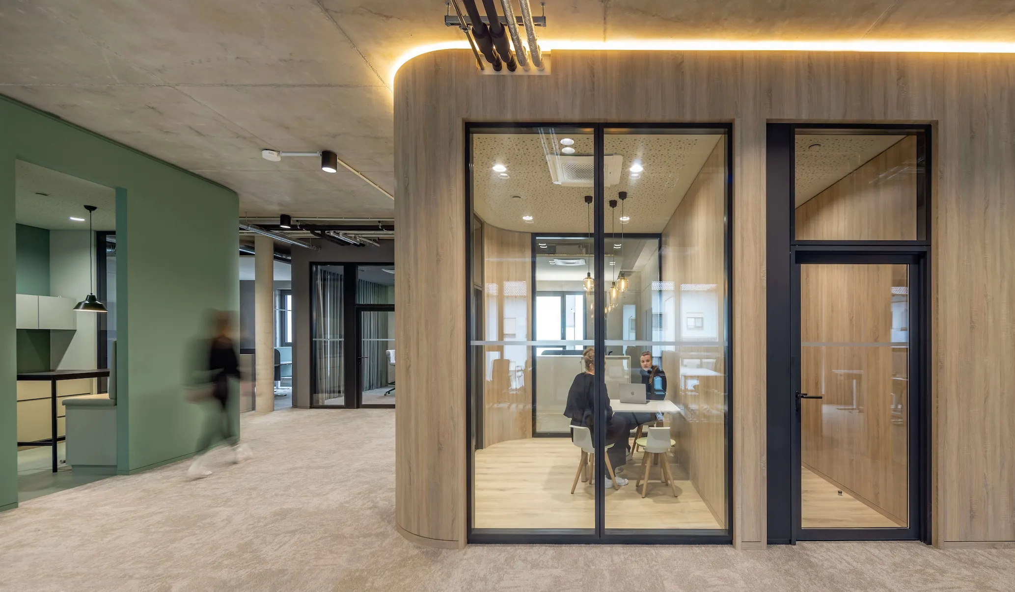 Modernes Büro mit verglastem Besprechungspod aus Holz: Zwei Personen im vertraulichen Meeting am Laptop, im Hintergrund offene Bürofläche mit grünen Akzenten.
