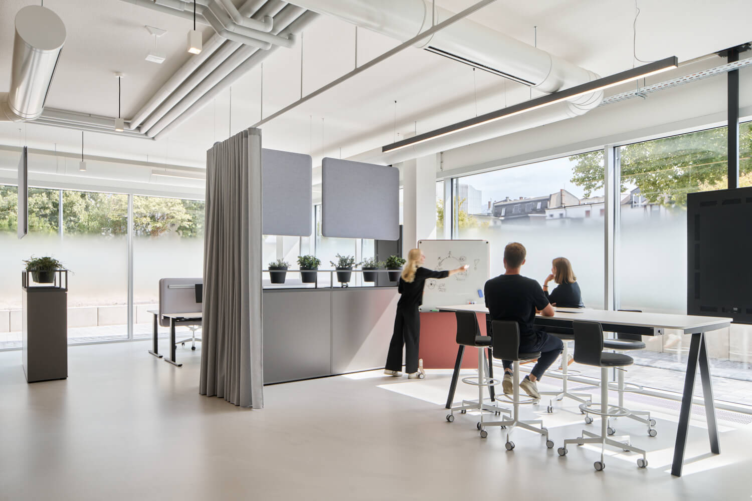 Helles, flexibles Büro mit Stehpulten und Whiteboard: Frau präsentiert zwei Kolleginnen und Kollegen in einer modernen, lichtdurchfluteten Arbeitszone mit Vorhang-Raumteilern und Grünpflanzen.