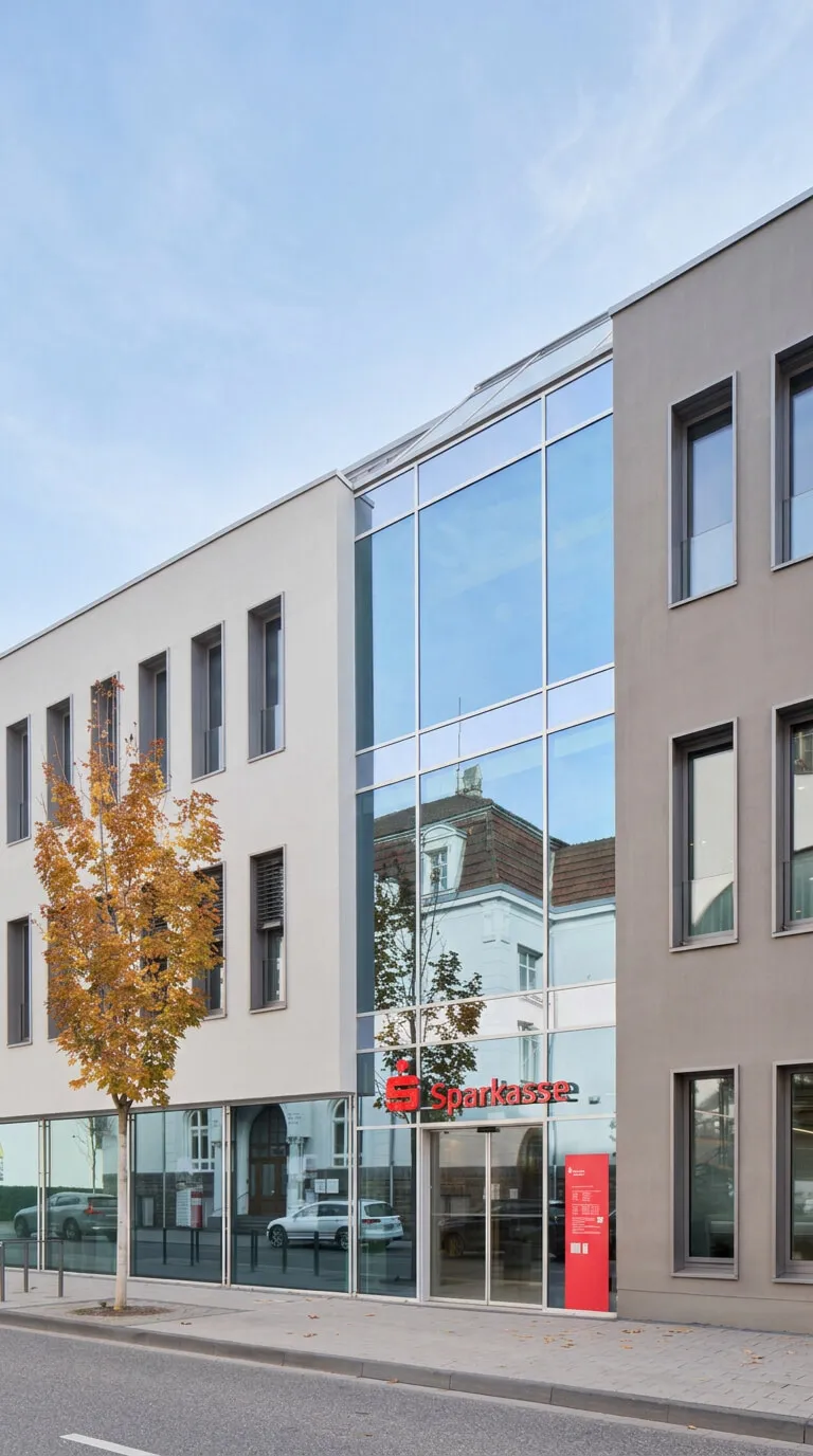 Außenansicht der Sparkasse-Filiale mit moderner Glasfront, rotem Sparkasse-Logo und herbstlichem Straßenbaum im Vordergrund.
