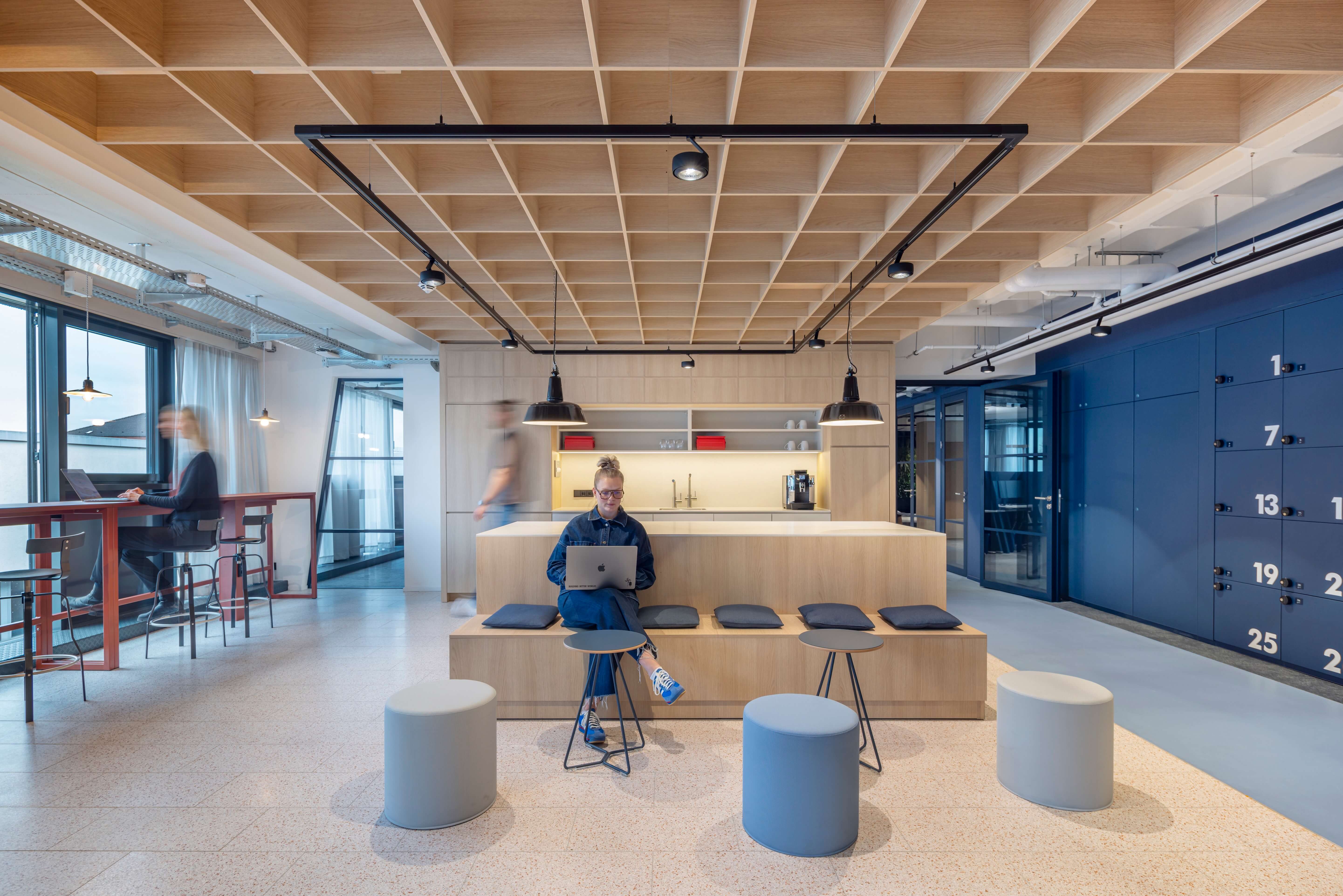 Moderner Büro-Sitz-Bereich mit Holztheke, integrierter Küchenzeile, blauen Schließfächern und strukturierter Holzdecke. Eine Frau arbeitet am Laptop an der zentralen Sitztheke.