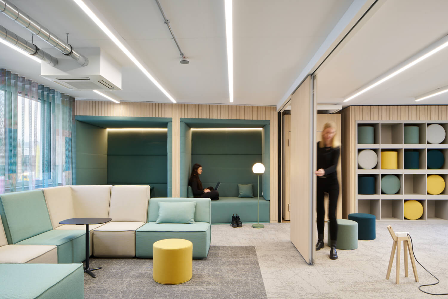 Flexibler Lounge- und Rückzugsbereich eines modernen Büros mit mintfarbenen Sofas, integrierten Nischen zum konzentrierten Arbeiten und einem offenen Regal mit bunten Sitzmöbeln – Innenarchitektur, die verschiedene Arbeitsmodi in einem Raum vereint.