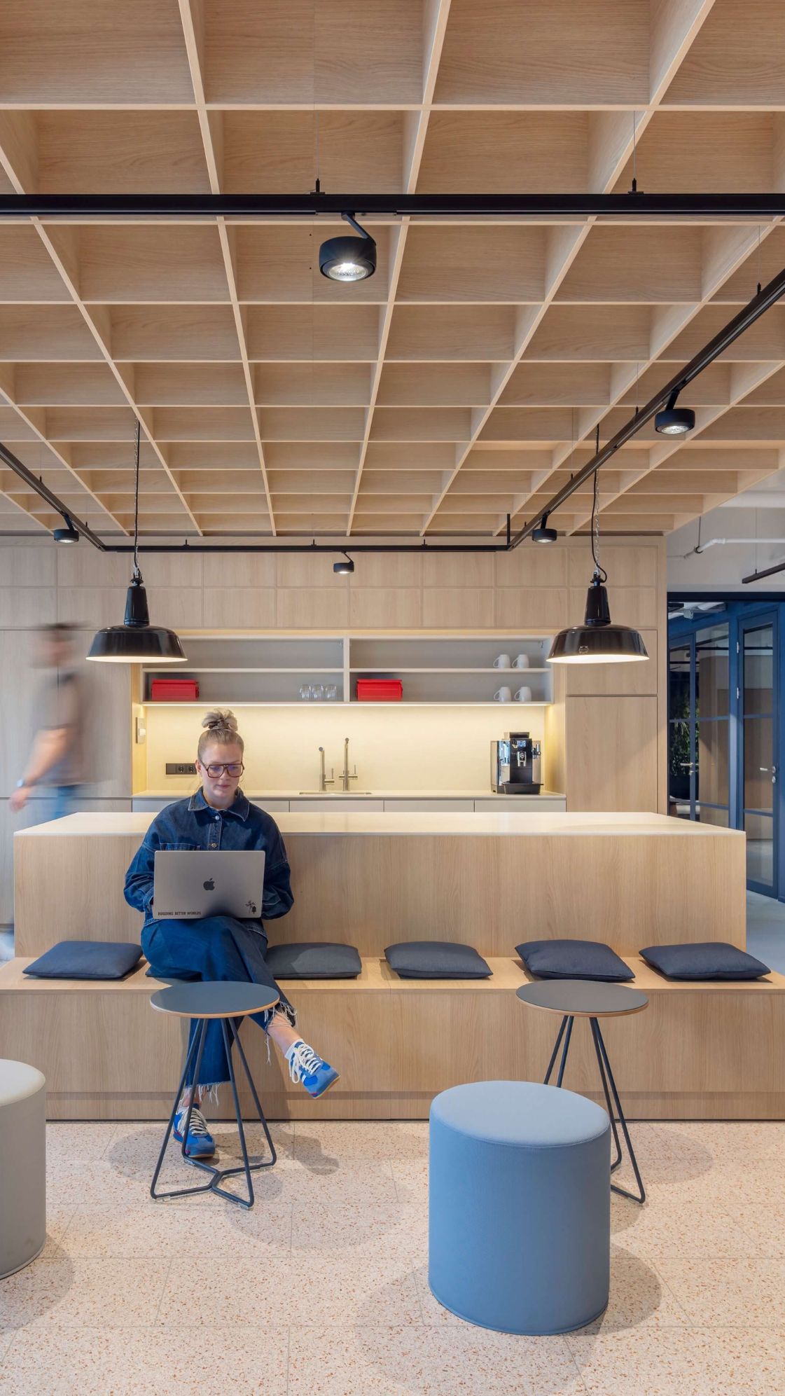 Moderner Büro-Sitz-Bereich mit Holztheke, integrierter Küchenzeile, blauen Schließfächern und strukturierter Holzdecke. Eine Frau arbeitet am Laptop an der zentralen Sitztheke.