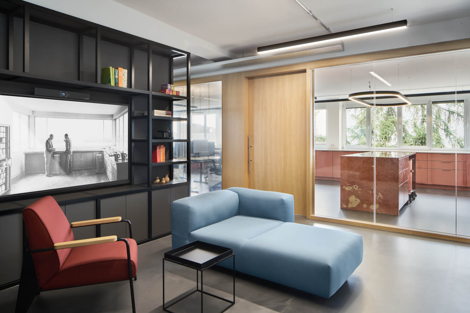 Stilvoller Lounge-Bereich eines modernen Büros mit blauem Sofa, rotem Sessel und dunklem Regalwandsystem – im Hintergrund öffnet sich der Raum zu einer eleganten Küche mit Marmor-Kücheninsel.