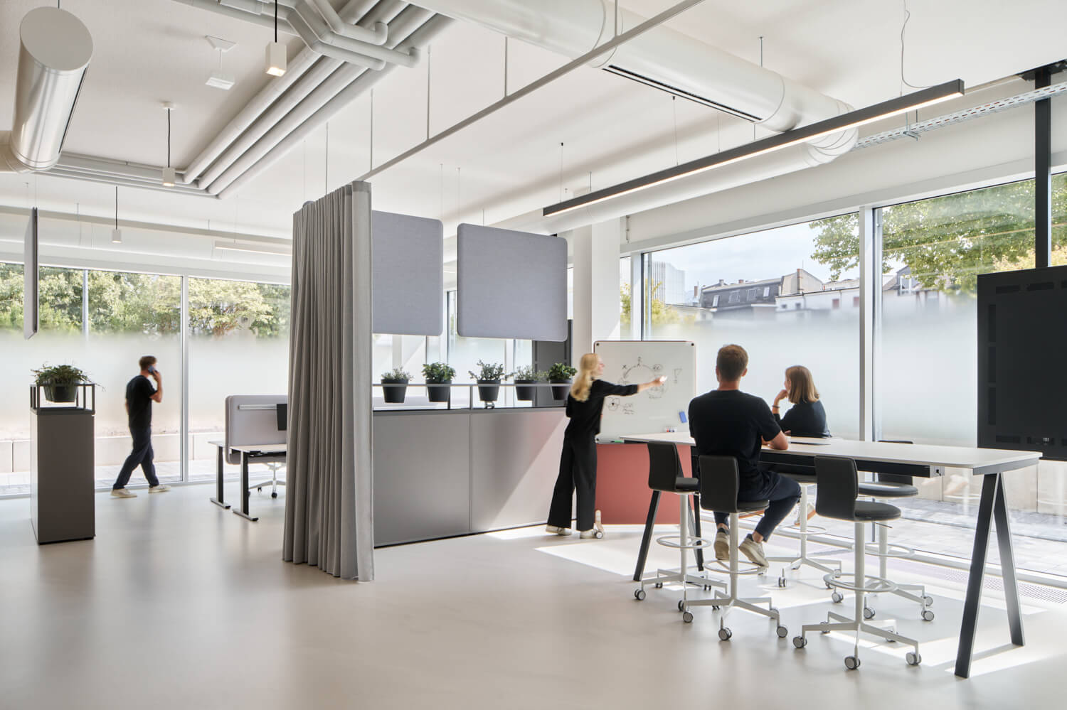 Modernes Büro mit flexiblen Arbeitsbereichen: Eine Mitarbeiterin erklärt am Whiteboard, während zwei Kollegen an einem Stehtisch zuhören – lichtdurchflutet mit Raumteilern und flexiblen Arbeitsmöglichkeiten.