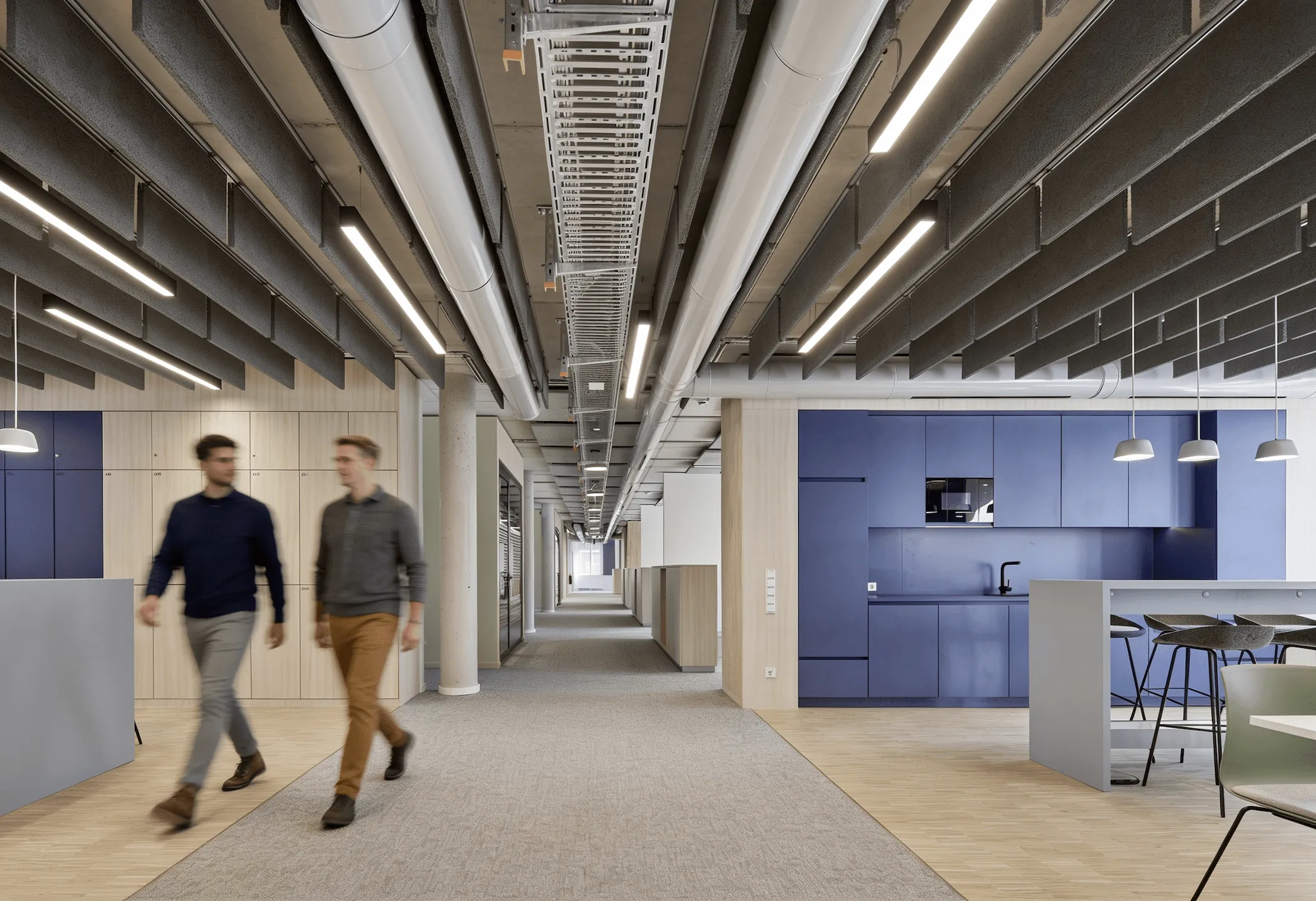 Moderner Büroflur mit Teppichboden, blauen Küchenmöbeln rechts und beleuchteten Deckenpaneelen.
