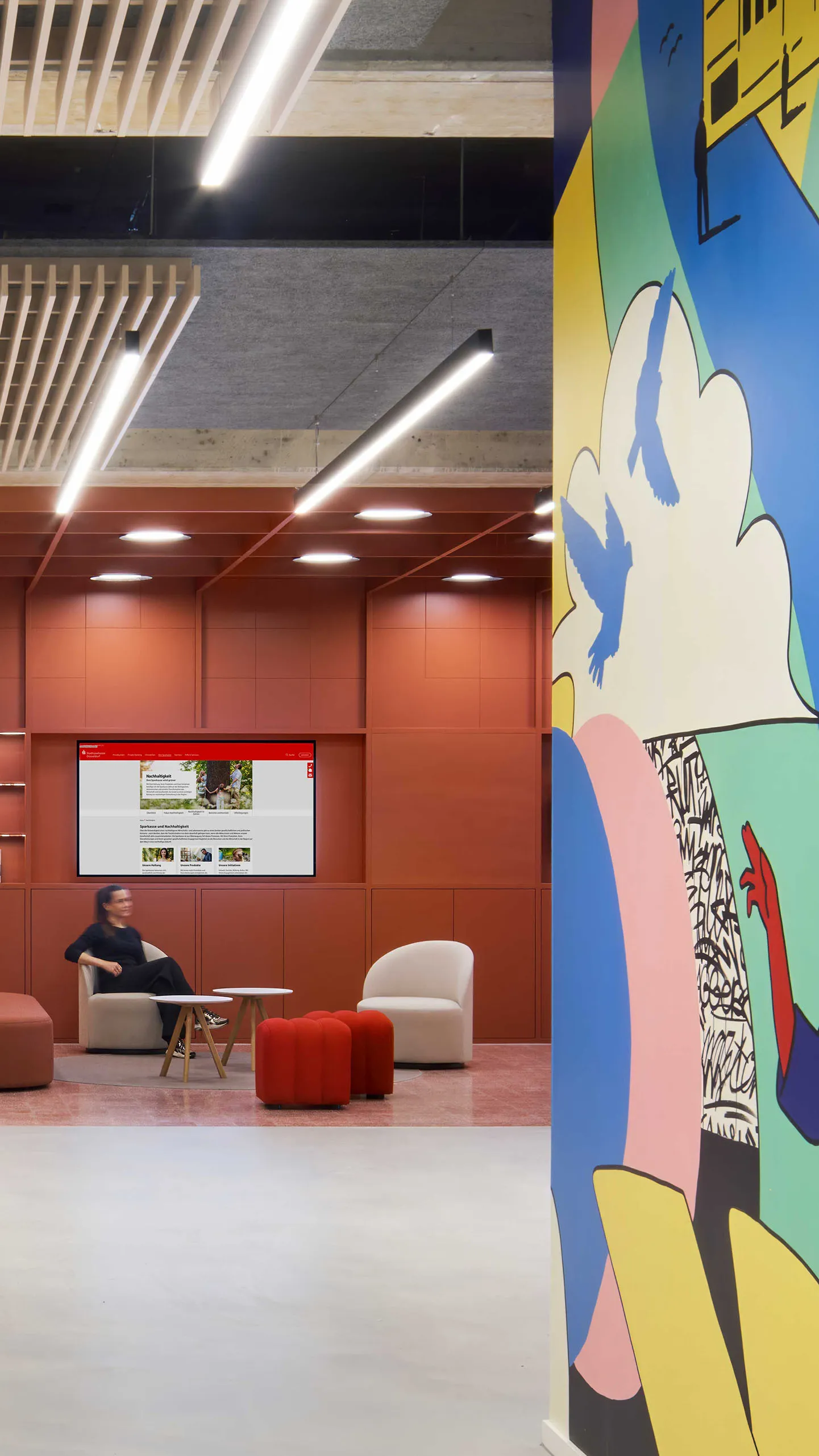 Moderner Büro Empfang mit hellem Holzmöbeln, Betonstütze, neonbeleuchtetem Sparkassen-Firmenlogo und bunter Wandgestaltung.