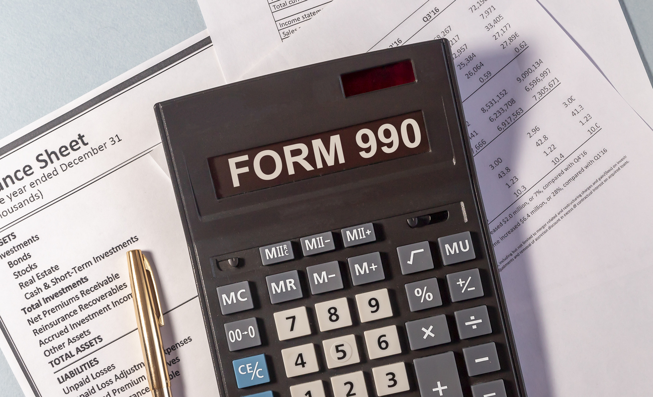 A calculator sits on top of accounting paperwork. The screen on the calculator reads “Form 990”