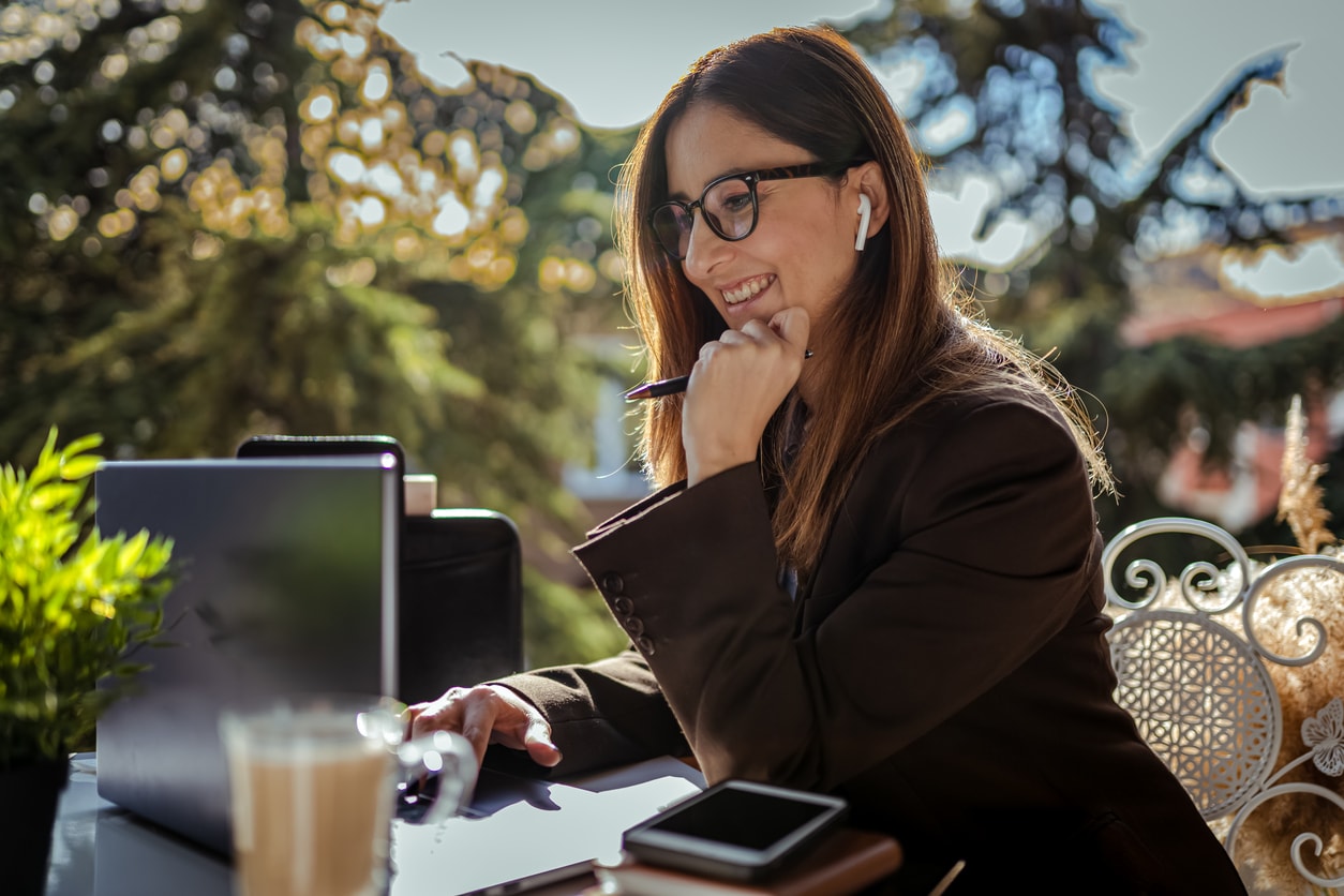 A photo of a woman smiling, working on a laptop and listening through earbuds. 