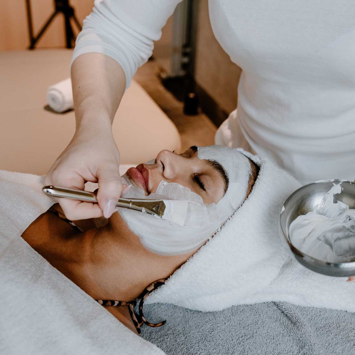 Frau erhält eine Wellness-Gesichtsbehandlung, während eine Person eine Gesichtsmaske mit einem Pinsel aufträgt.