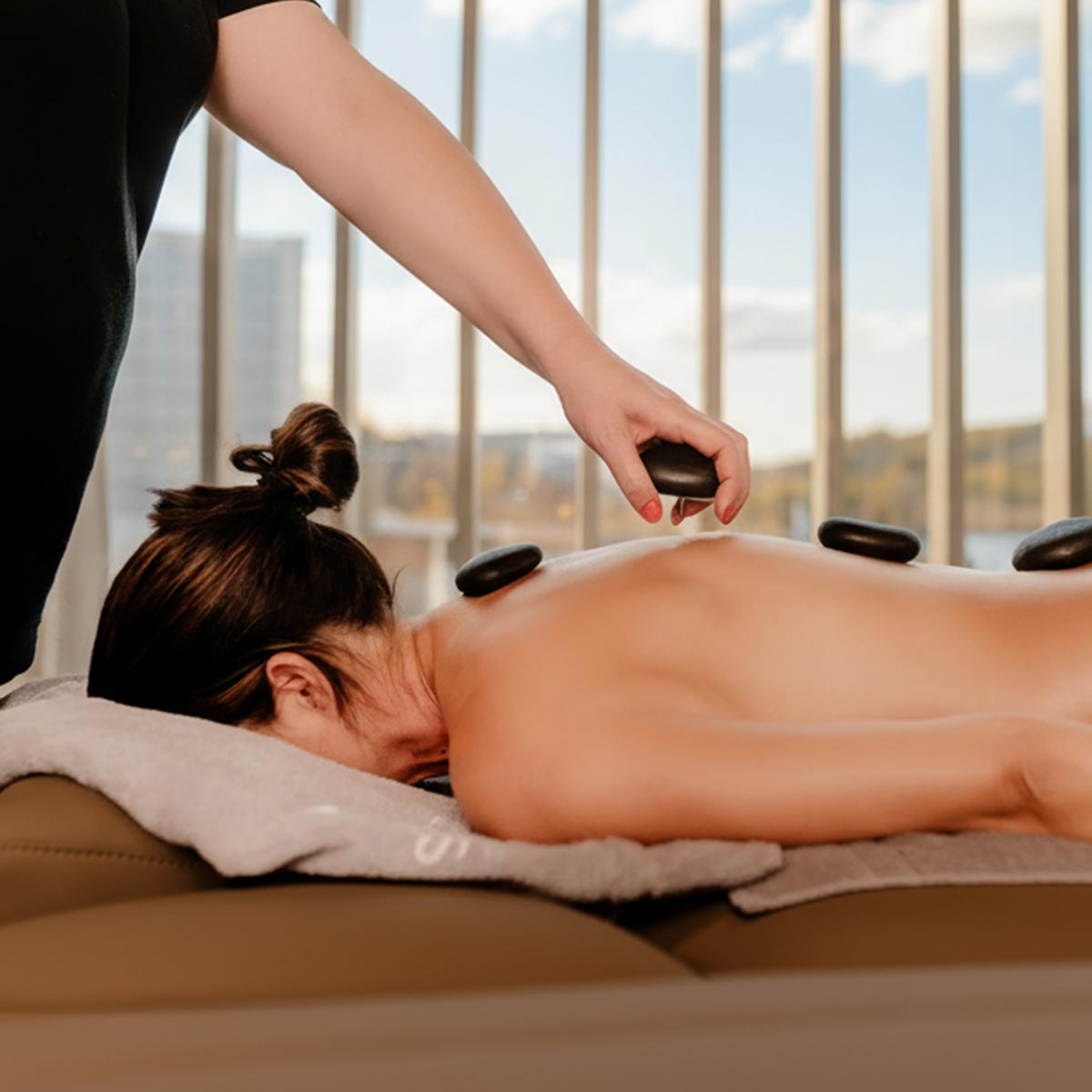 Eine Person erhält eine Hot-Stone-Massage mit schwarzen Steinen auf dem Rücken in einem hellen Raum mit großen Fenstern im Wellness-Bereich der Salinarium Therme Bad Dürkheim