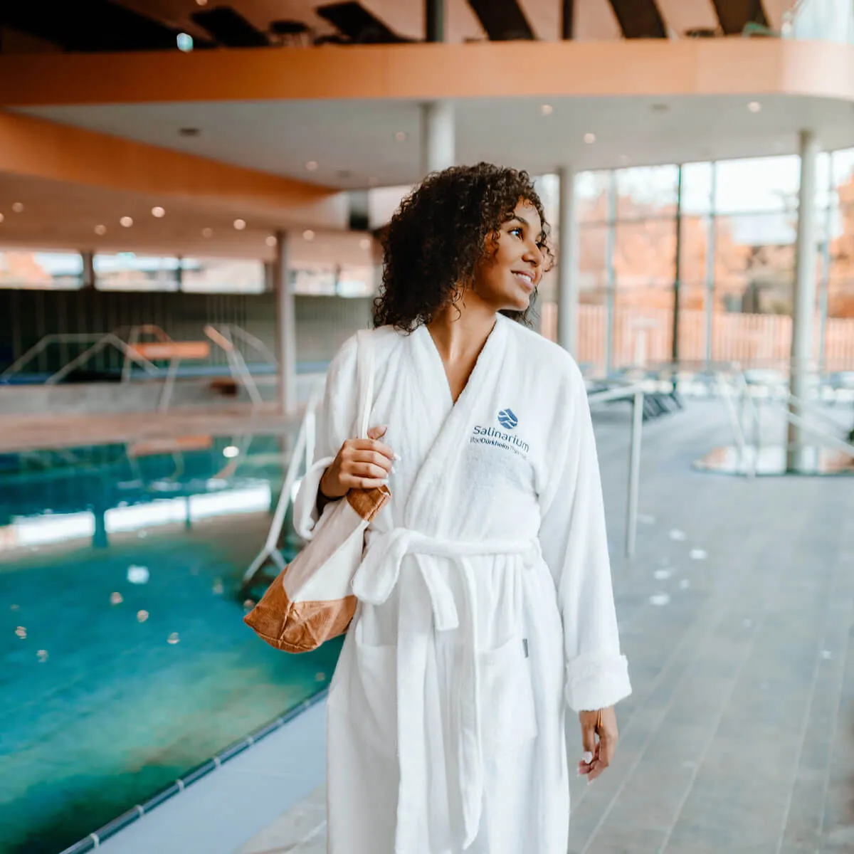 Frau in weißem Bademantel mit Tasche steht im Thermalbadbereich der Salinarium Bad Dürkheim Therme.