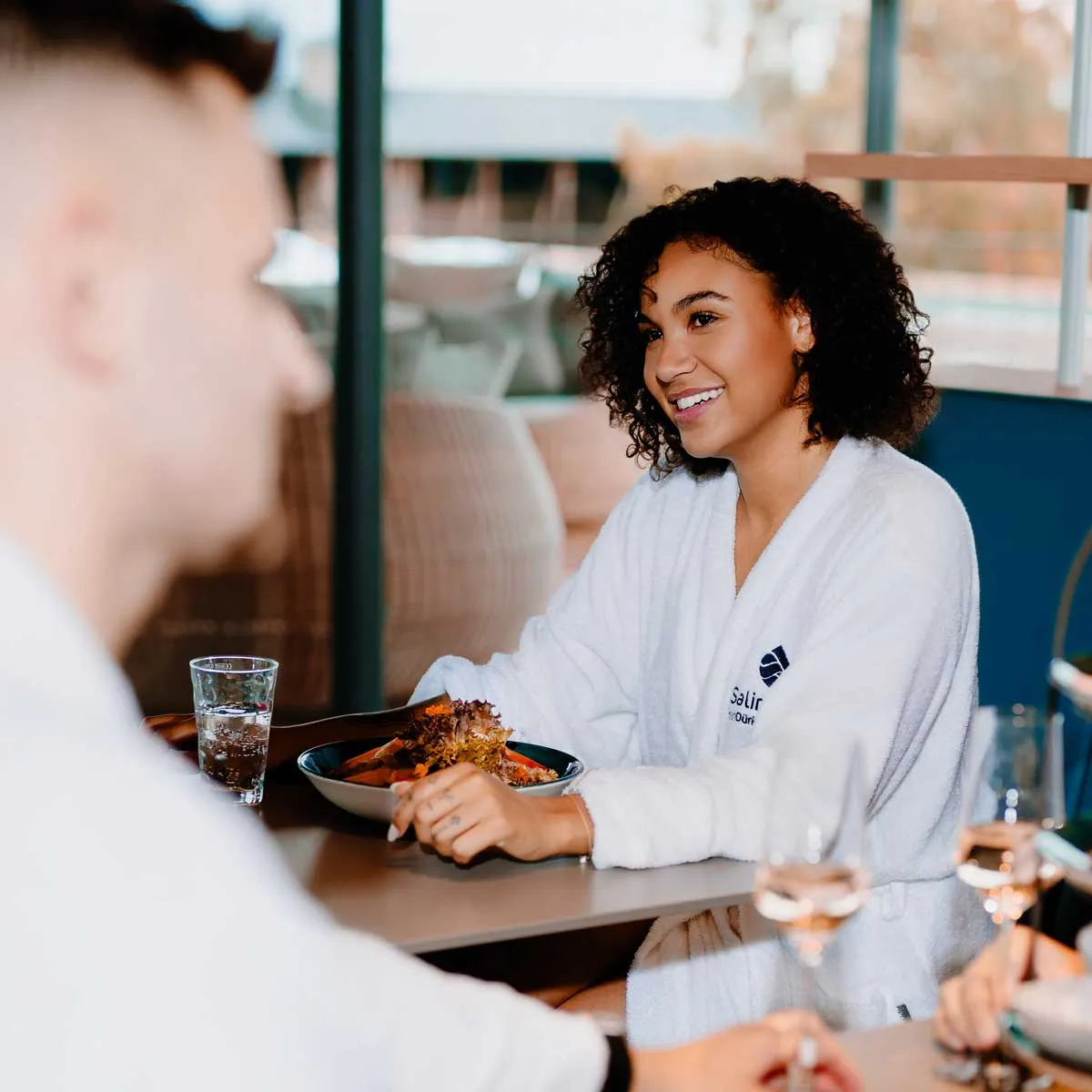 Frau in weißem Bademantel sitzt lächelnd bei einem Tisch mit Salat und Getränken, während sie mit anderen spricht in der Salinarium Bad Dürkheim Therme