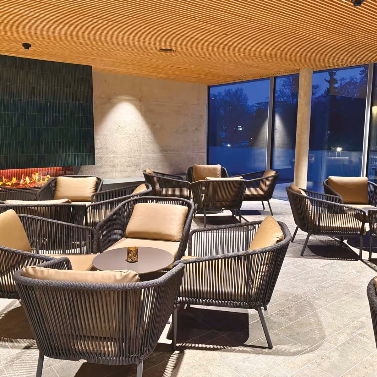 Moderne Lounge mit gepolsterten Stühlen um Tische, Holzdeckenpaneele und einem Kamin an einer Betonwand bei Abenddämmerung.