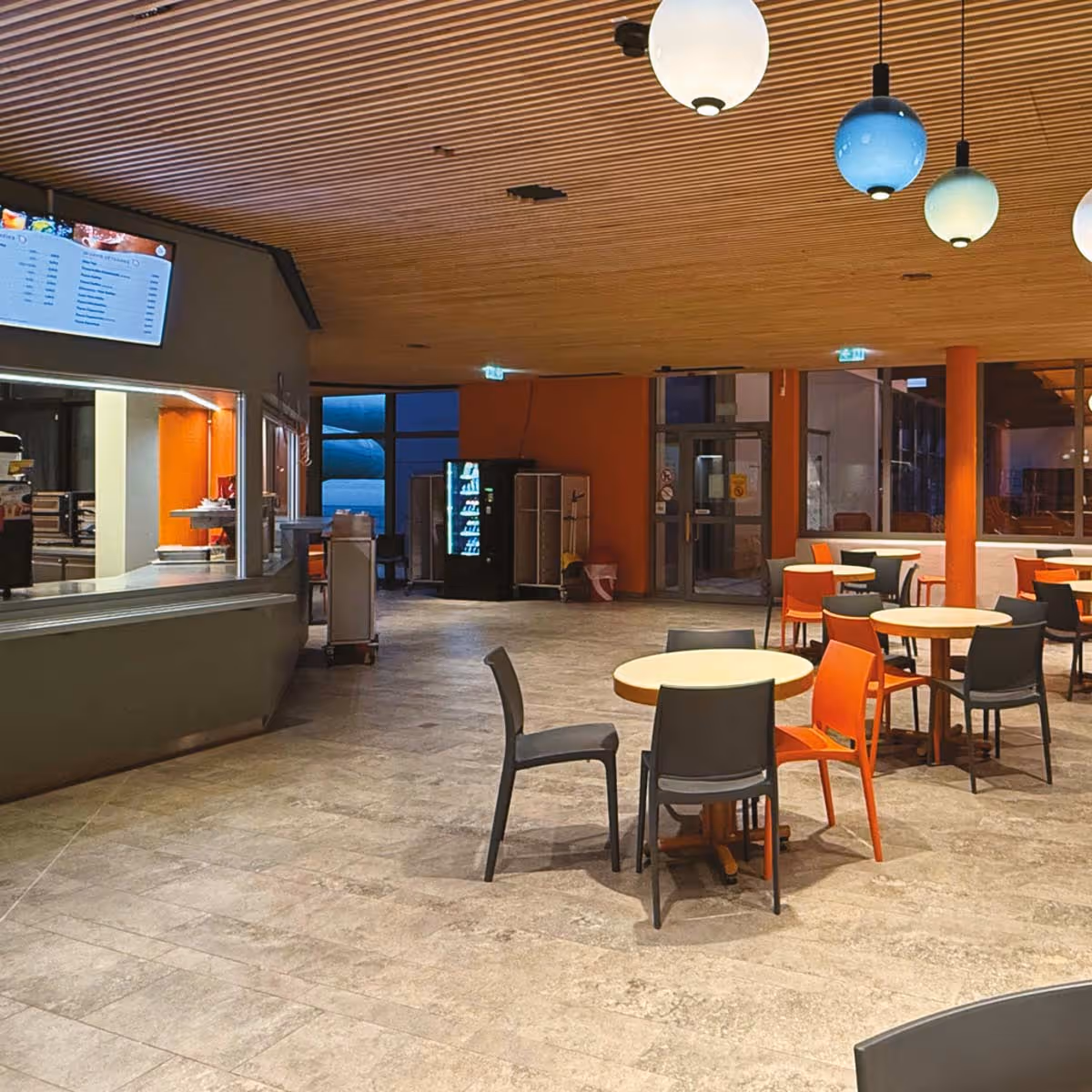 Moderne Cafeteria mit runden Tischen, orangenen und grauen Stühlen und einer Servicetheke mit digitalem Menübildschirm.