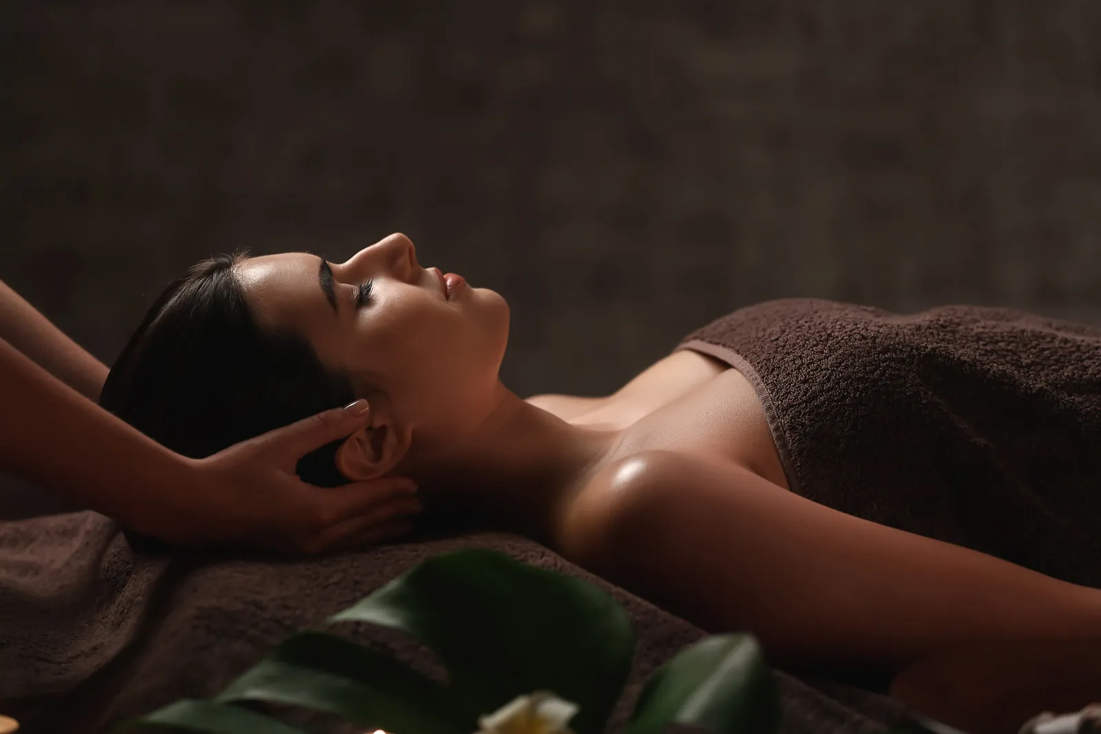 Frau entspannt liegend mit geschlossenen Augen, während eine Massage am Kopf im gedämpften Wellness-Umfeld durchgeführt wird.