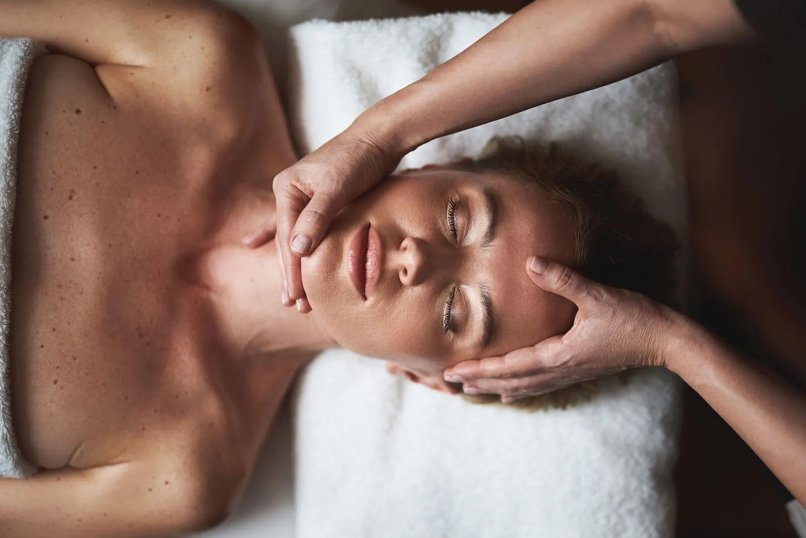 Frau entspannt mit geschlossenen Augen while sie eine Gesichtsmassage auf einer weißen Liege erhält.