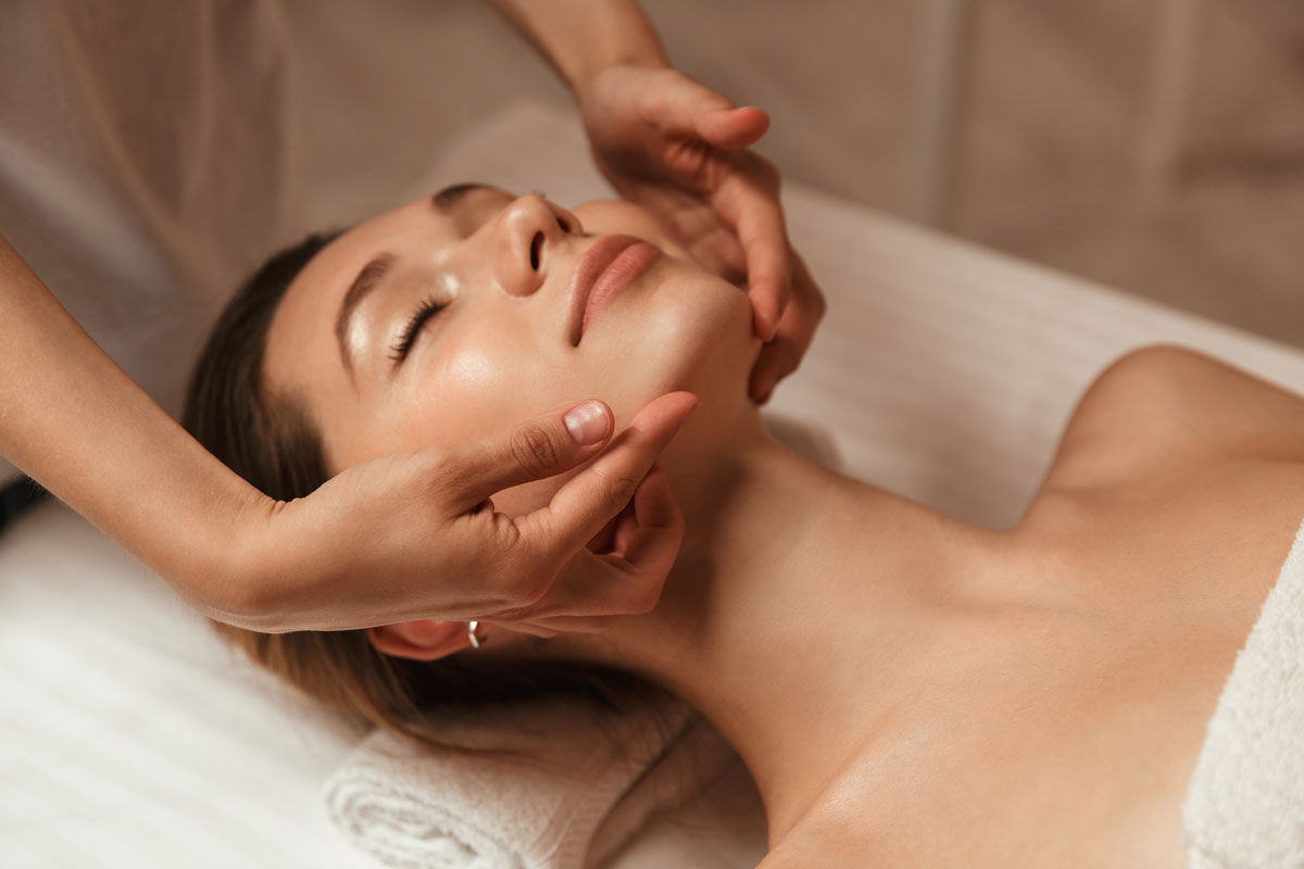 Frau mit geschlossenen Augen erhält Gesichtsmassage, liegt entspannt auf einer weißen Liege mit Handtuch.