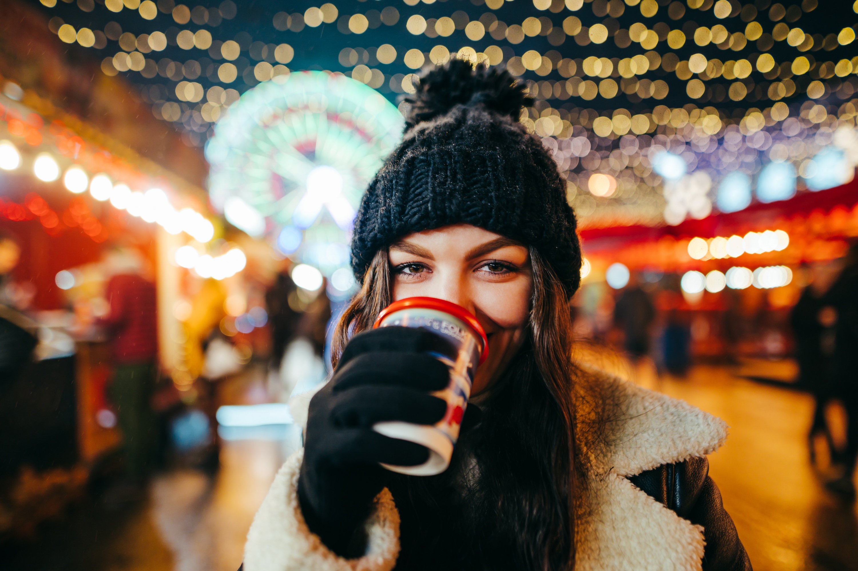 A resident smiling and holding a hot drink while wearing a knit hat and gloves at an illuminated outdoor night market or event, illustrating nearby community attractions and local events.