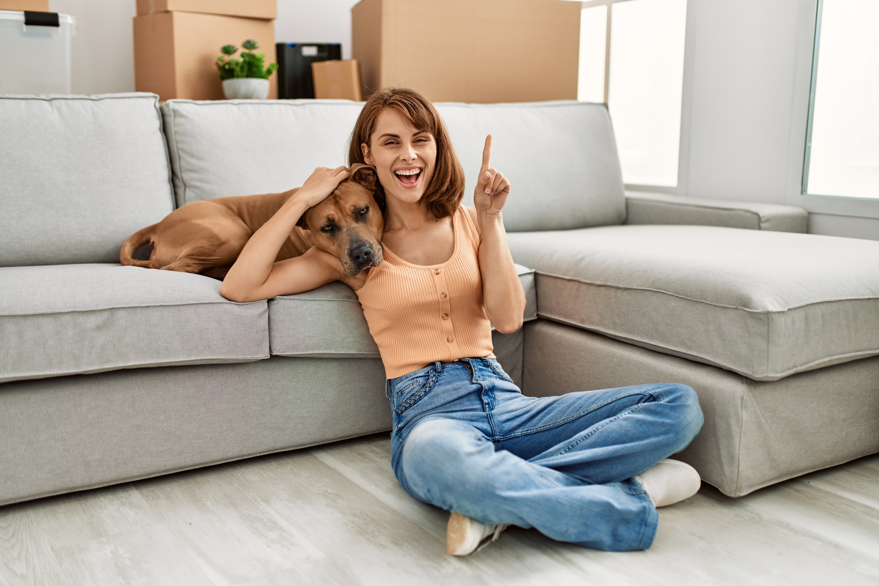 A happy resident sitting on the floor with their dog in a spacious living room at Wildcat Vista, featuring modern gray sofas and moving boxes in the background to showcase a pet-friendly move-in experience.