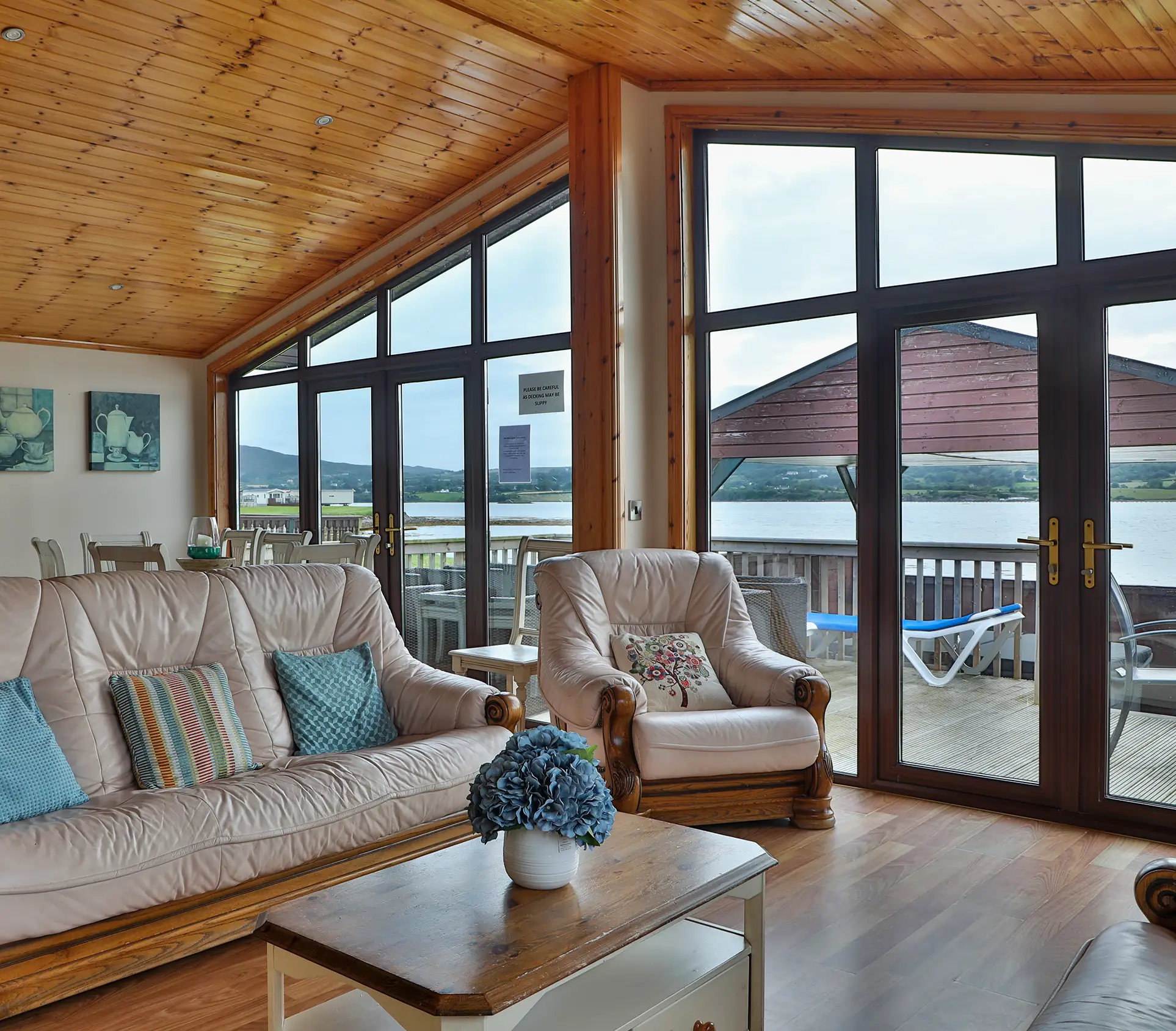 Cozy living room with leather sofas, coffee table, and large windows overlooking a lake and deck.