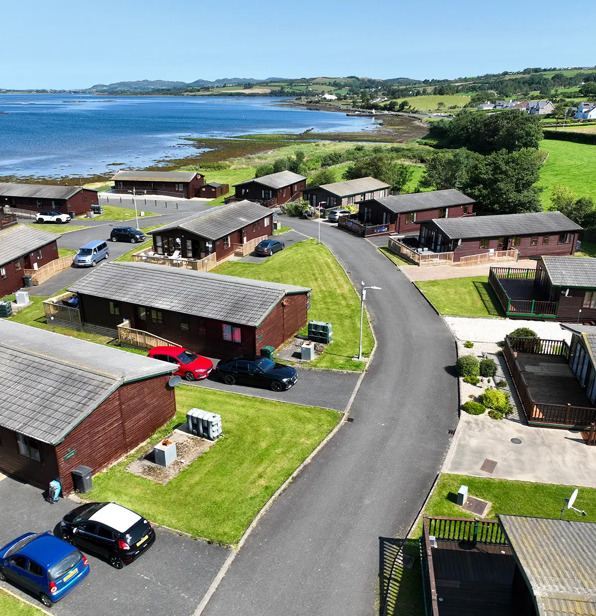 Aerial view of lakeside cabins with parked cars, green lawns, a curved road, and hills in the background.