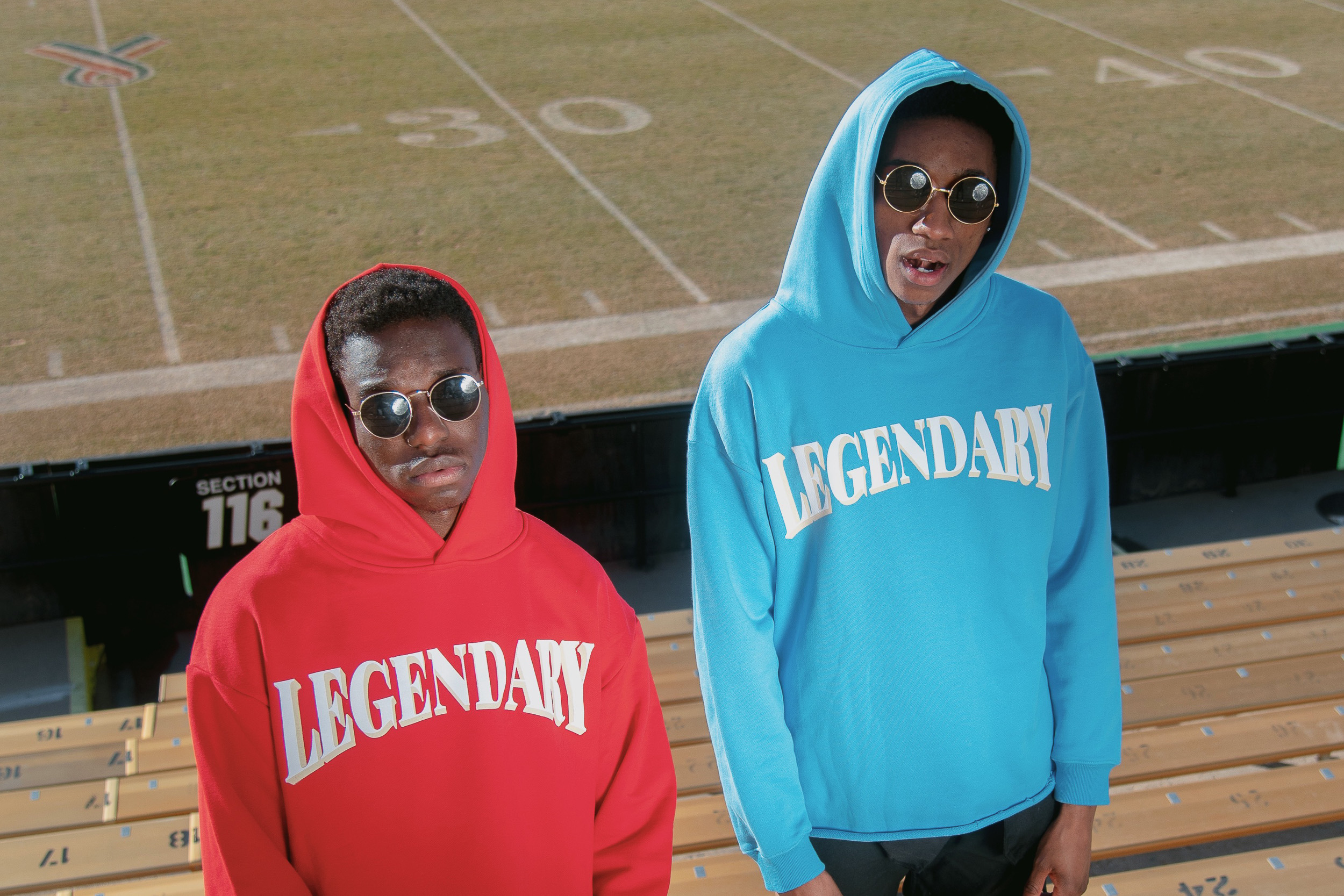 Two models in red and blue Legendary hoodies standing on stadium bleachers overlooking the field