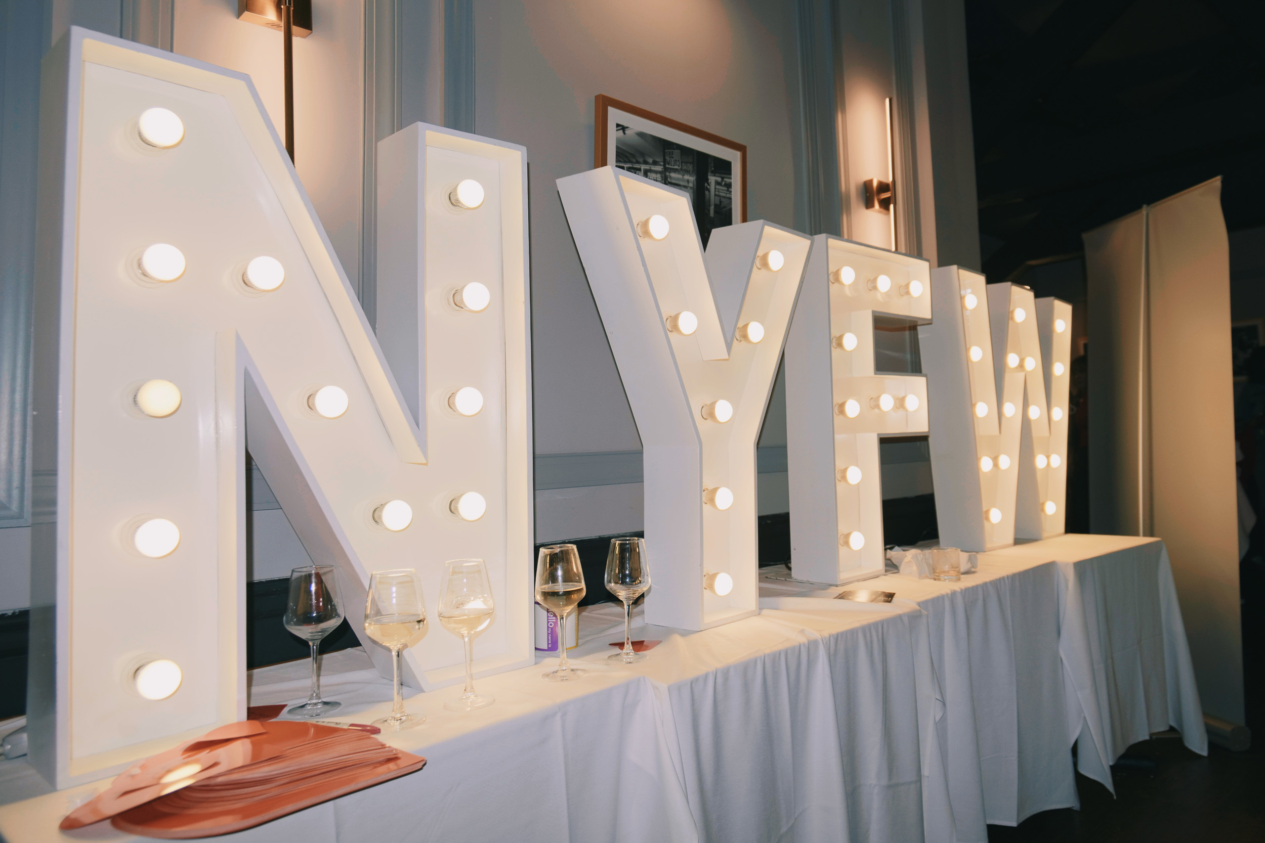 Lit NYFW marquee letters on a white event table with wine glasses