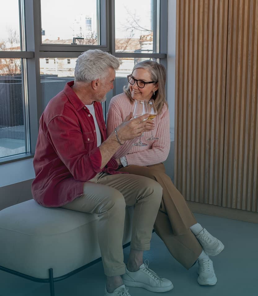 Older couple sitting indoors by large windows, smiling and clinking wine glasses in a toast.