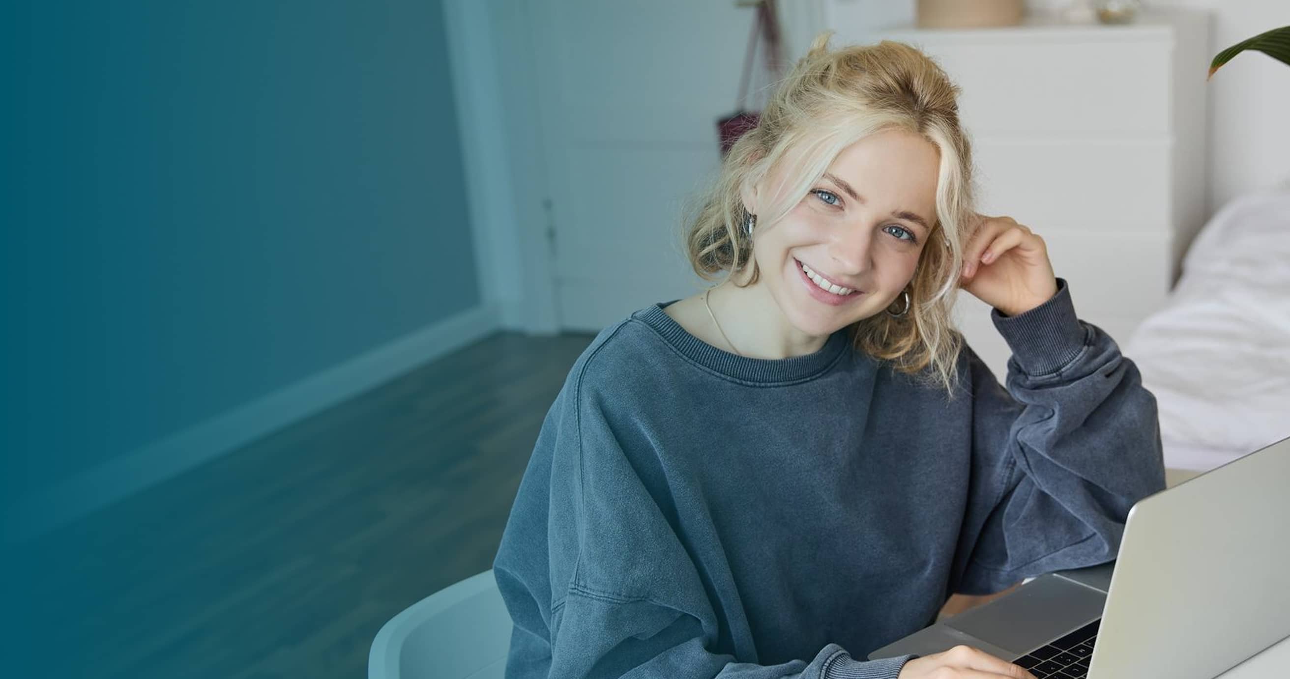 Smiling young woman with blonde hair wearing a gray sweatshirt sitting at a desk with a laptop.