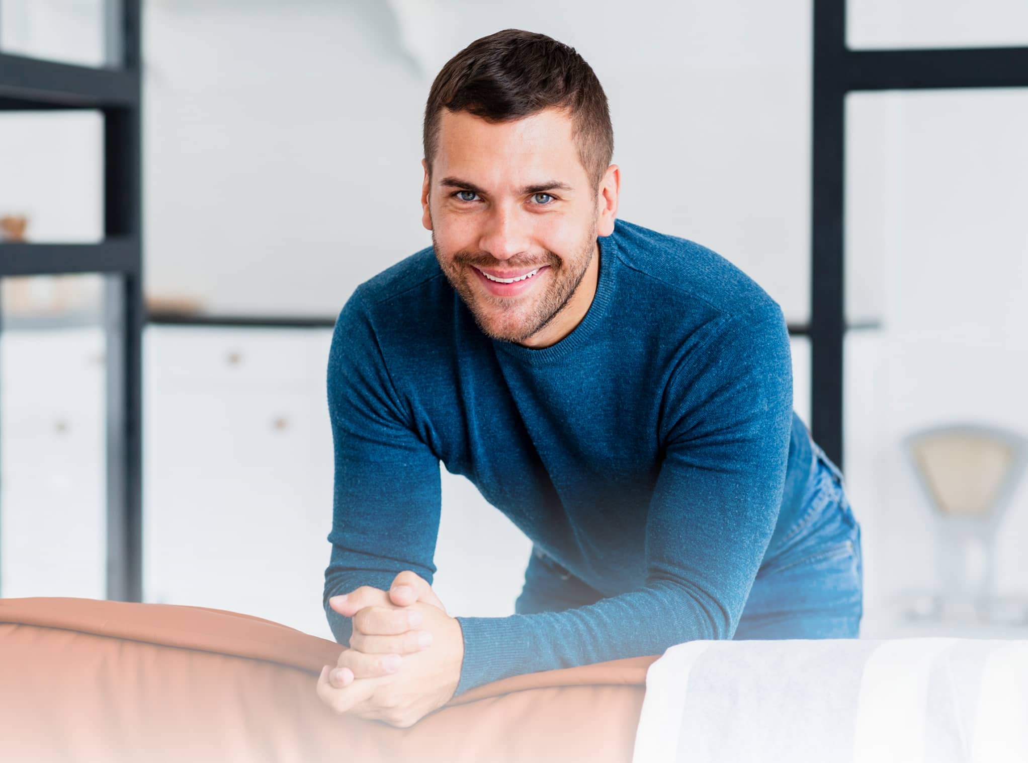 Smiling man in a blue sweater leaning forward with clasped hands on an orange couch in a bright room.