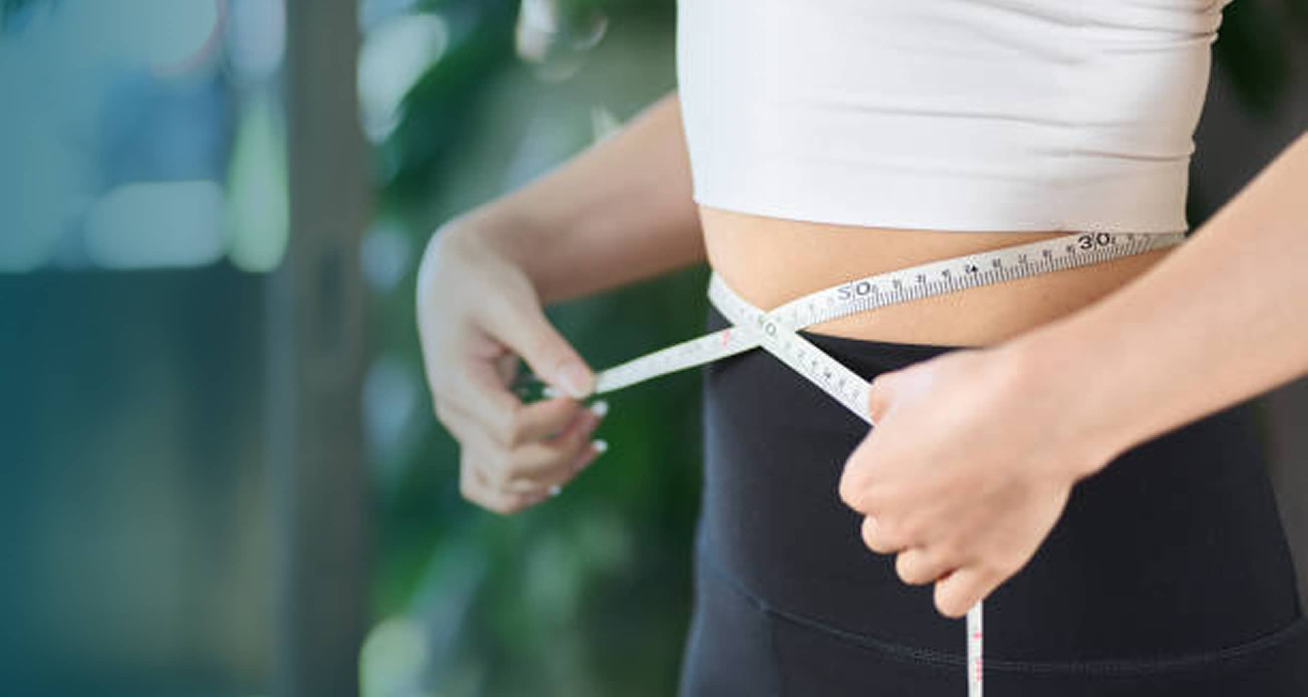 Person measuring their waist with a soft measuring tape while wearing a white top and black bottoms.