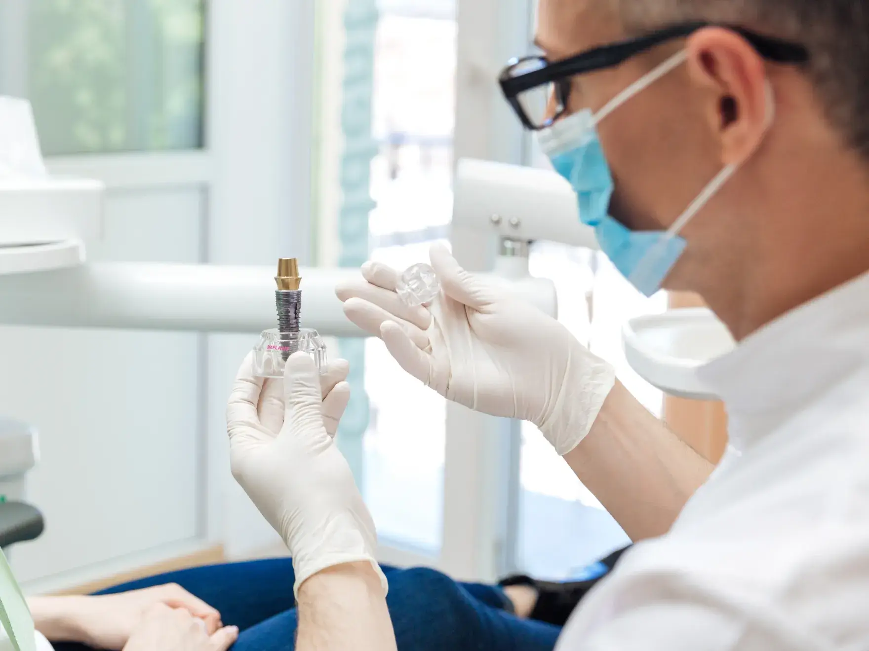 A dentist wearing gloves and a mask explains a dental implant to a patient in an office.
