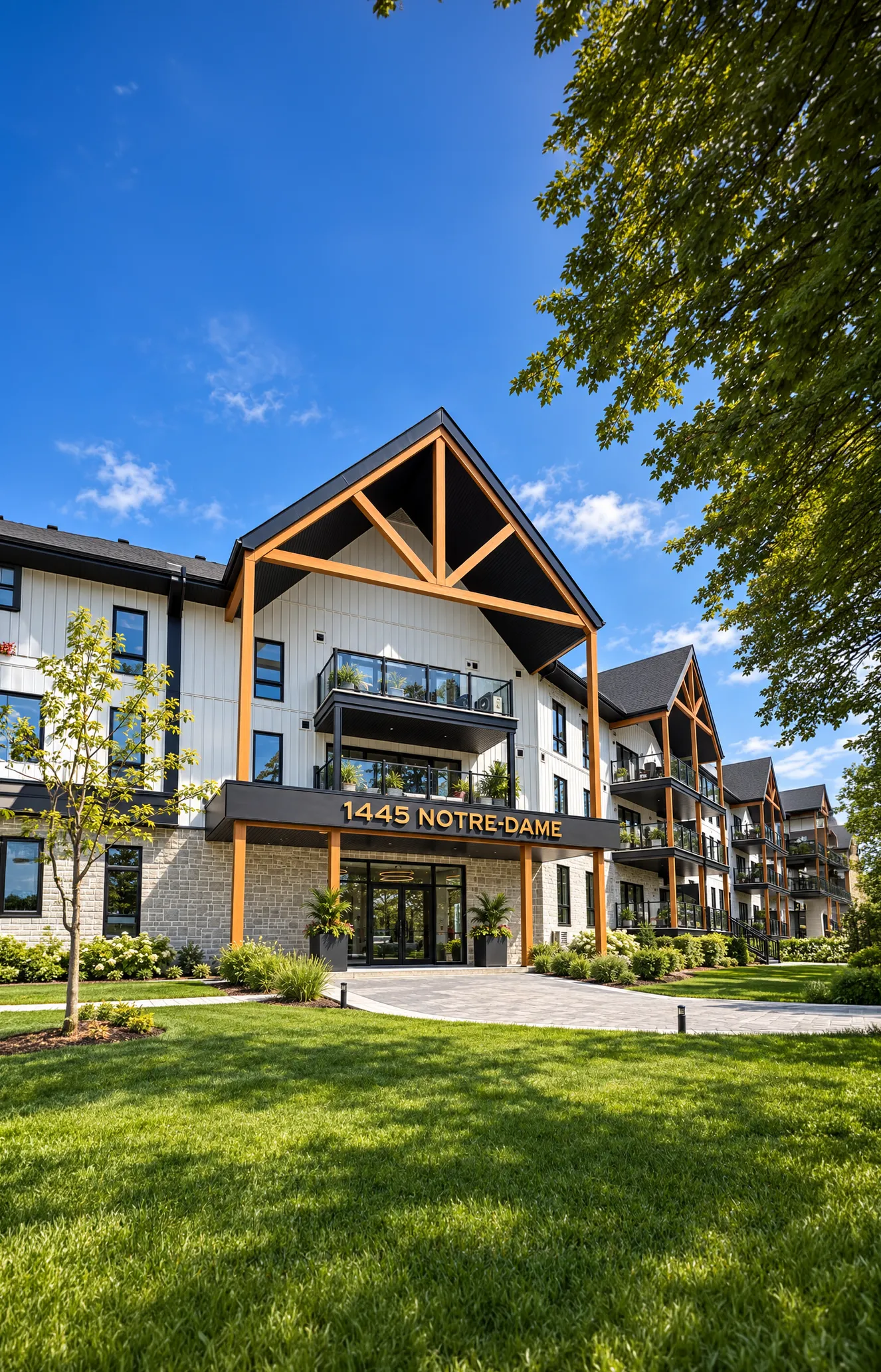 Modern multi-story residential building with balconies, white and stone facade, wooden beams, and the address 1445 Notre-Dame above the entrance, surrounded by green lawn and trees under a blue sky.