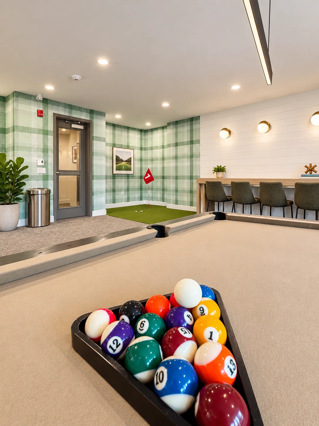 Indoor game room featuring a beige pool table with a full rack of colorful billiard balls in the foreground, a small indoor putting green with a red flag in the background, green plaid wallpaper on two walls, a door, a trash can, and a long wooden counter with four dark green chairs and round wall lights above it.