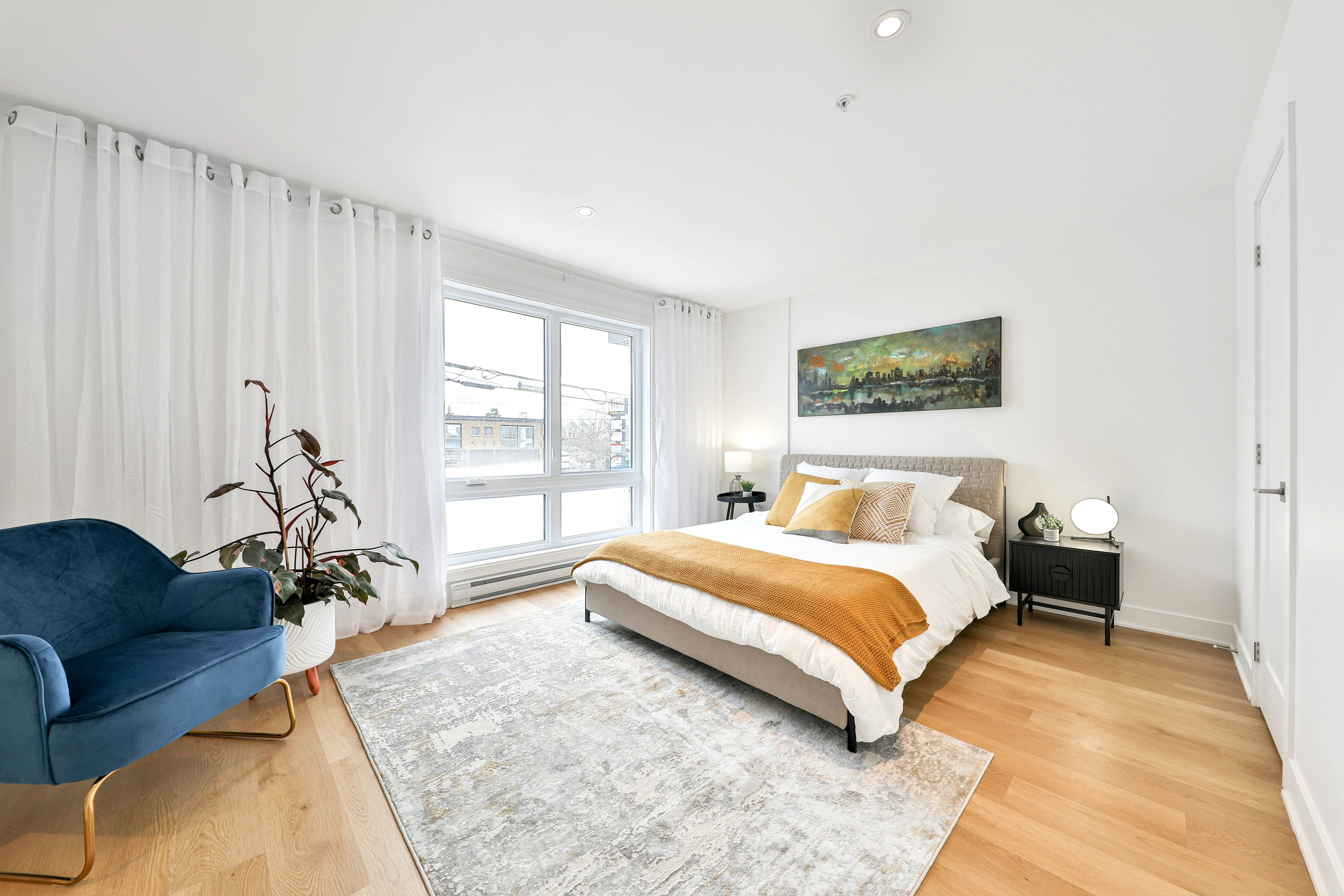 Bright modern bedroom with light wood floor, large window with sheer white curtains, gray upholstered bed with white bedding and mustard yellow throw and pillows, black nightstand with lamp and small decor, blue armchair, potted plant, and cityscape painting on white wall.
