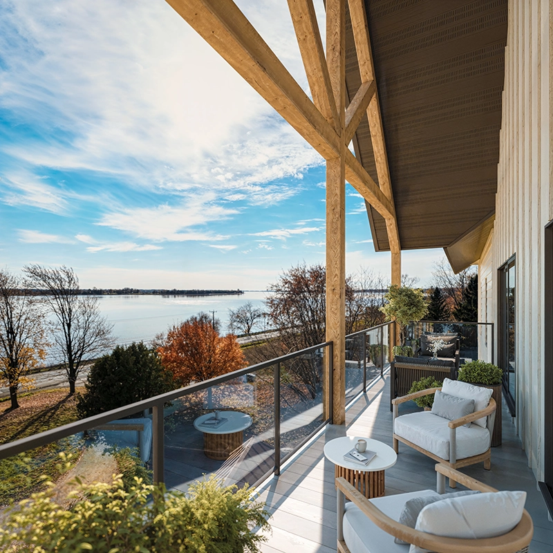 Spacious balcony with modern wooden furniture including cushioned chairs and round tables, overlooking a serene body of water and autumn trees under a partly cloudy sky.