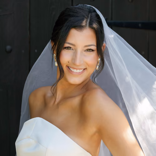 A close-up portrait of a smiling bride wearing a white strapless gown, a sheer veil, and pearl drop earrings.