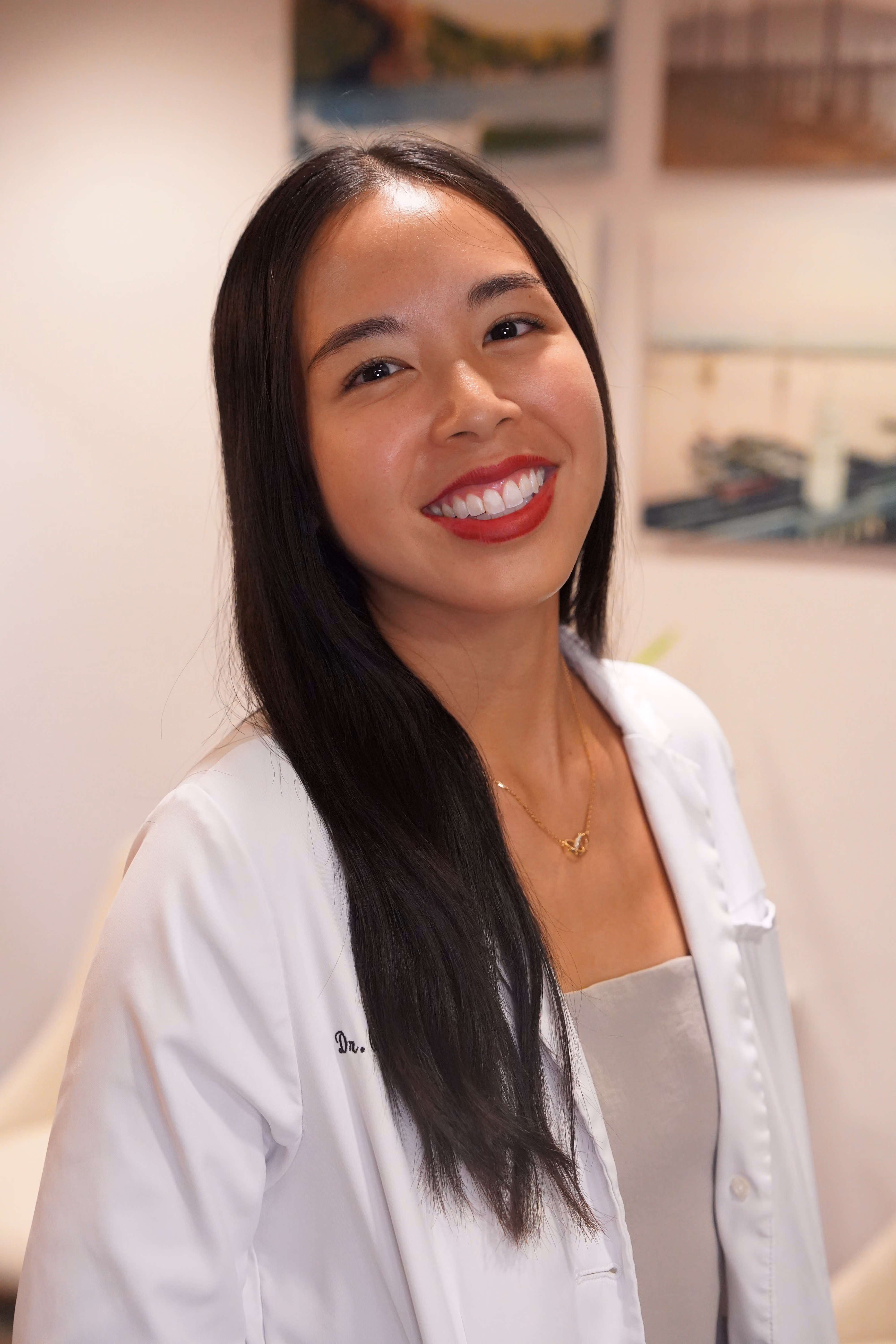 Smiling woman with long dark hair in white coat and red lipstick in an indoor setting.